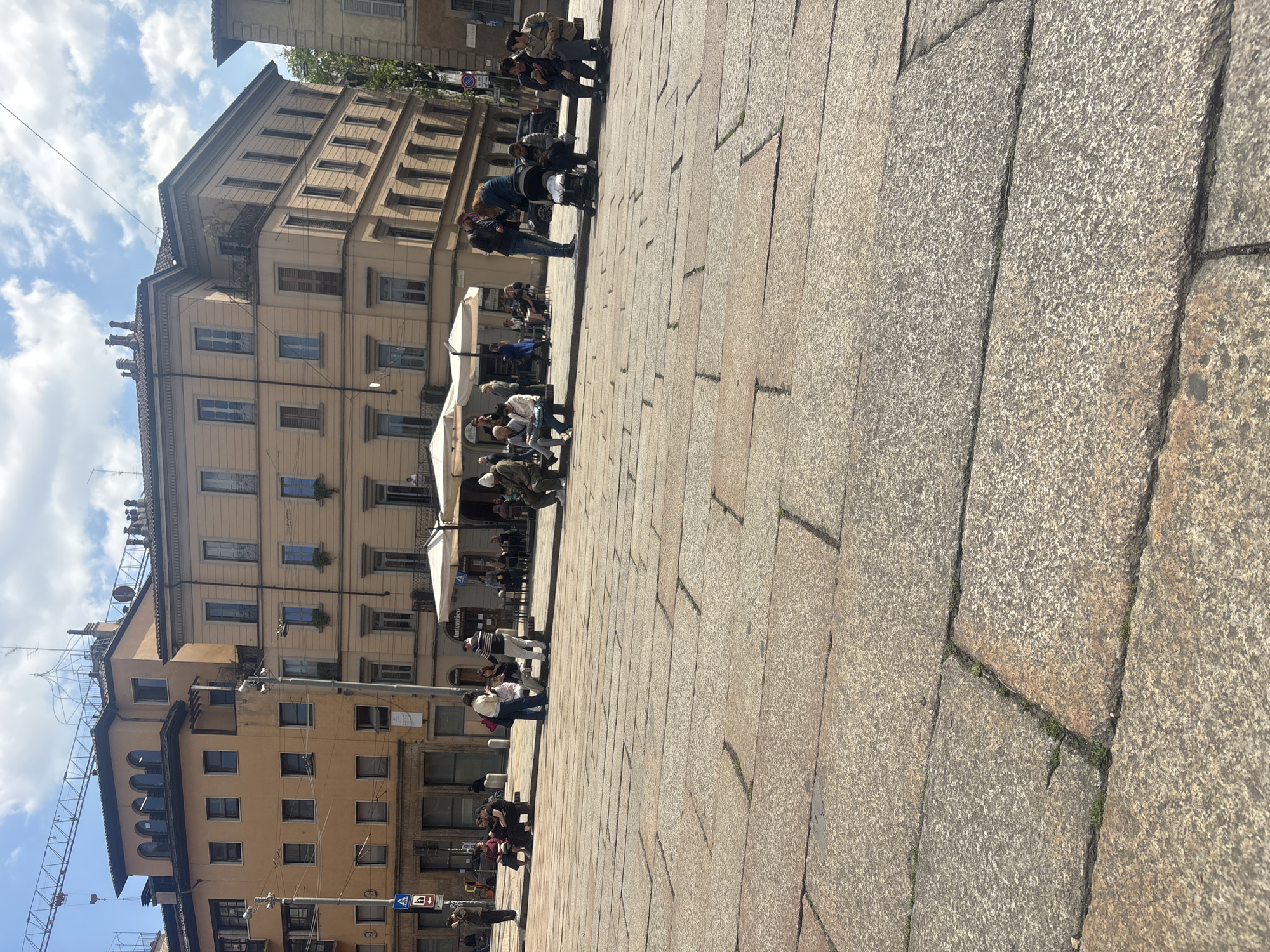 This daytime image captures a bustling urban square or wide pedestrian street in Milan, Italy. The scene is dominated by a large, textured stone-paved ground extending from the foreground into the mid-distance, and multi-story buildings lining the left side of the frame.
The paving consists of large, irregular granite slabs, showing signs of wear and small patches of grass or moss growing in the cracks. Overhead, the sky is bright blue with scattered cumulus clouds, indicating sunny weather, likely during the mid-day or afternoon, as evidenced by the distinct shadows cast across the ground.
On the left, a row of architecturally distinct buildings rises several stories high. These structures feature numerous windows, many equipped with dark-colored shutters, and some include small iron balconies adorned with planters. Extending from the ground level of these buildings are several white awnings or large umbrellas, providing shade for outdoor seating areas, suggesting cafes or restaurants.
Various individuals and small groups of people are visible throughout the space. They are casually dressed and engaged in typical urban activities: walking, standing, sitting at the outdoor establishments, and conversing. In the mid-ground, a cluster of people is gathered near a lamppost, while others are distributed further back.
Notable objects include a tall street light pole with several luminaires, and a network of overhead wires stretching across the sky. Several traffic signs are visible, including a blue circular sign with a white right-pointing arrow (indicating a mandatory turn), and another sign further back that appears to be a no-parking or no-stopping sign (red circle with a diagonal line). Although some text is faintly visible on one of the white awnings, it is too small and obscured to be clearly legible. The overall atmosphere is lively and typical of a European city center.