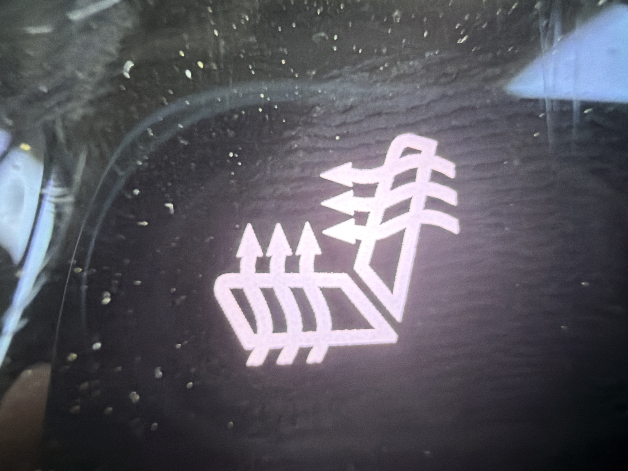 The image shows a close-up, illuminated icon on a dark surface, likely a car's interior dashboard. The icon is a stylized representation of a car seat with arrows indicating airflow. Three upward-pointing arrows are shown emanating from the bottom of the seat, and two arrows point towards the top of the seat. This icon is universally recognized as a symbol for ventilated seats, indicating a feature that circulates air through the seat to cool the occupant. The lighting suggests this is likely an indoor environment, possibly a car dealership or a private garage, and the time of day is indeterminate. There is no visible text in the image. The surface the icon is on appears to be slightly dusty or textured.