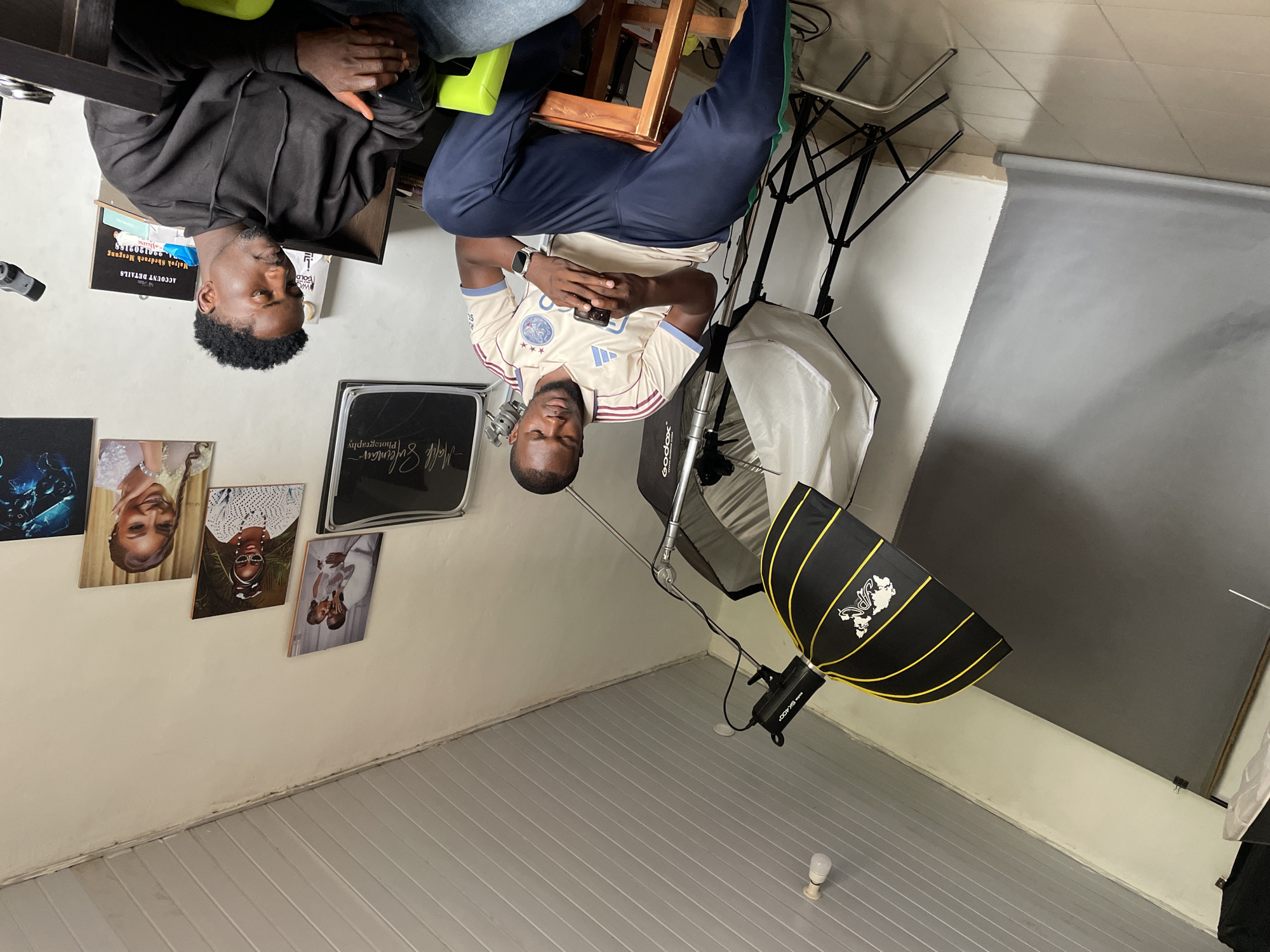 The image shows two men in what appears to be a photography studio or a room set up for creative work. The first man, on the left, is wearing a black hoodie and is seated, with his hands clasped. A document with text is visible near him. The second man, on the right, is wearing a light-colored jersey with a design on it and is also seated, looking down with his hands clasped. He is wearing a smartwatch.
The room contains photographic equipment, including lighting stands, softboxes, and a large gray backdrop. Several framed pictures or prints are displayed on the wall, featuring portraits of people. The flooring is light-colored and appears to be wood or laminate. A small lightbulb is visible on the floor in the lower right portion of the image. The overall impression is a creative space, possibly a studio where photoshoots take place. The location is indicated as Jos, Nigeria. The lighting suggests an indoor environment, and there are no obvious cues about the time of day or weather. The emotions of the subjects are neutral, with both men appearing relaxed.
Visible text includes "ACCOUNT DETAILS", "MATSIYAH GBOCHOKO MEUNG", and "AWKO GOLD". There is also text on the jersey of the man on the right, but it is not legible. The title on one of the framed pictures reads "Mals Sulenman — Photography".