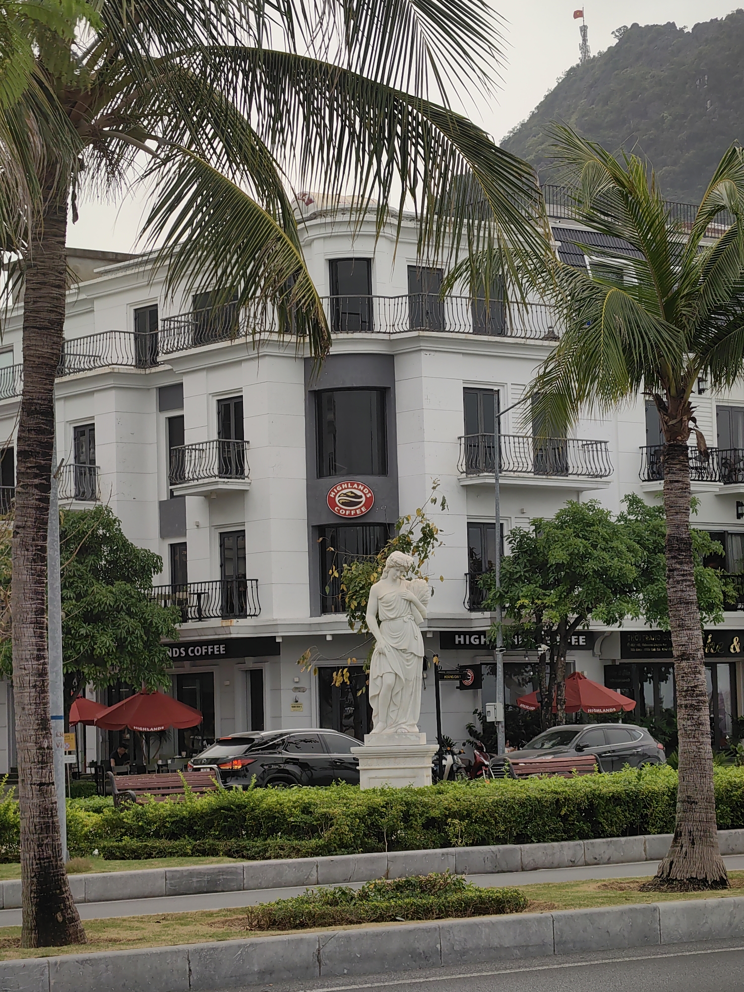 The image depicts a three-story building in Hong Gai Ward, Vietnam. The building is predominantly white with black accents, including balconies and window frames. A "Highlands Coffee" sign is prominently displayed. On the ground floor, the business name "Highlands Coffee" is visible above the entrance. In front of the building, a white statue stands on a pedestal surrounded by green shrubbery. Two palm trees frame the building. A road runs in front of the shrubbery, with two dark-colored cars parked along the side. Red umbrellas are visible outside the coffee shop, with some benches and a few people seated. A mountain is visible in the background, with a tower and a flag at the peak.