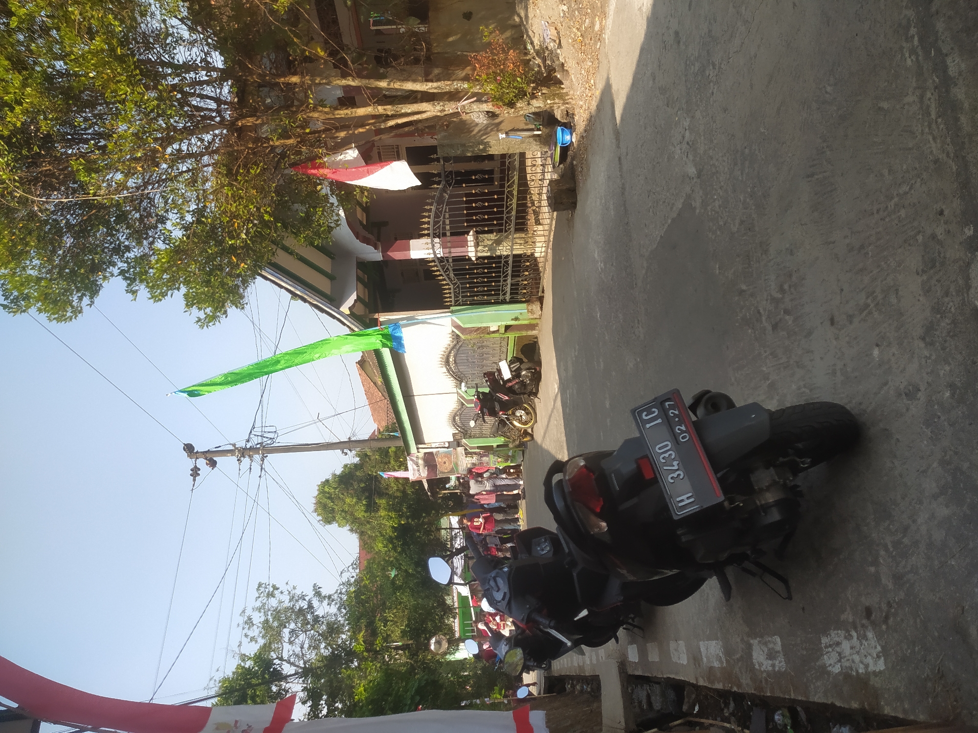 This is an outdoor, eye-level, vertical shot taken on a sunny day in Lopait, Indonesia. In the foreground, a black motorcycle is parked on a paved road. The motorcycle has a license plate that reads "H 3430 IC" and "02-27". Behind the motorcycle, several other motorcycles are parked along the side of the road. A group of people, possibly students, are standing in front of a building with a black wrought-iron fence. The building appears to be a house, with a red and white Indonesian flag hanging from a pole. A green banner is also visible, hanging from a telephone pole. The environment is residential, with trees and houses lining the street. The sky is clear and blue.