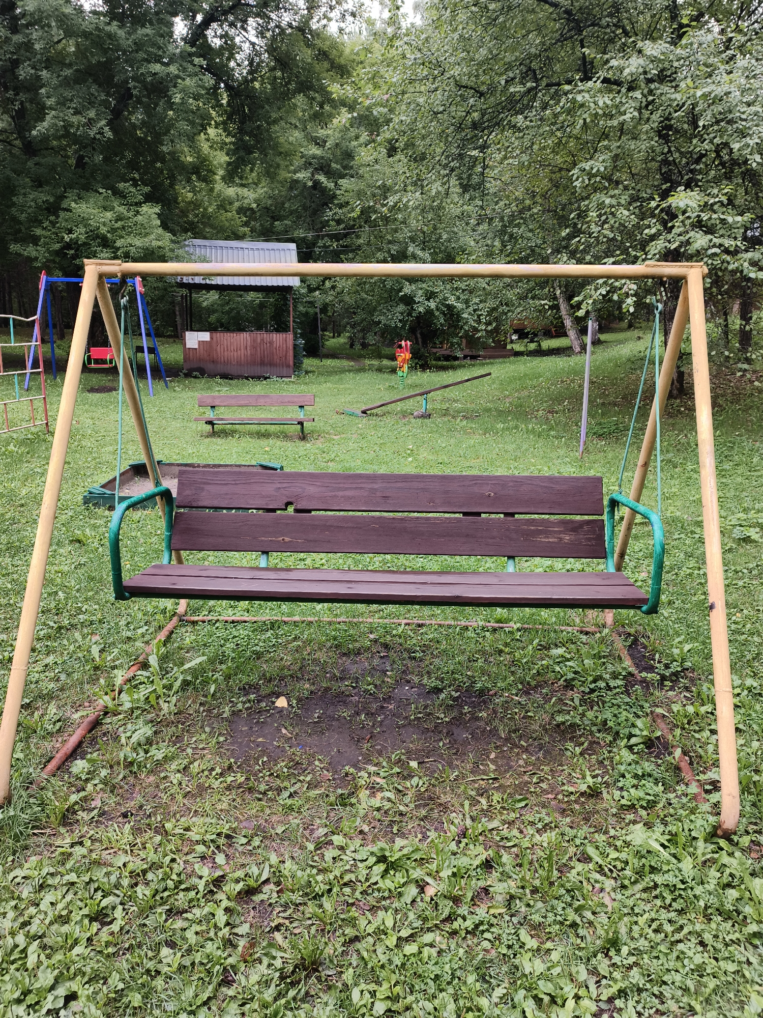 An outdoor scene in Катунь, Russia, features a playground with a wooden swing set in the foreground. The swing set has a yellow metal frame and a brown wooden bench with green metal armrests. Behind the swing set, there is a sandbox, a wooden bench, a seesaw, and other playground equipment, all situated on a grassy area surrounded by trees. The sky is overcast, and the overall atmosphere is calm and deserted.