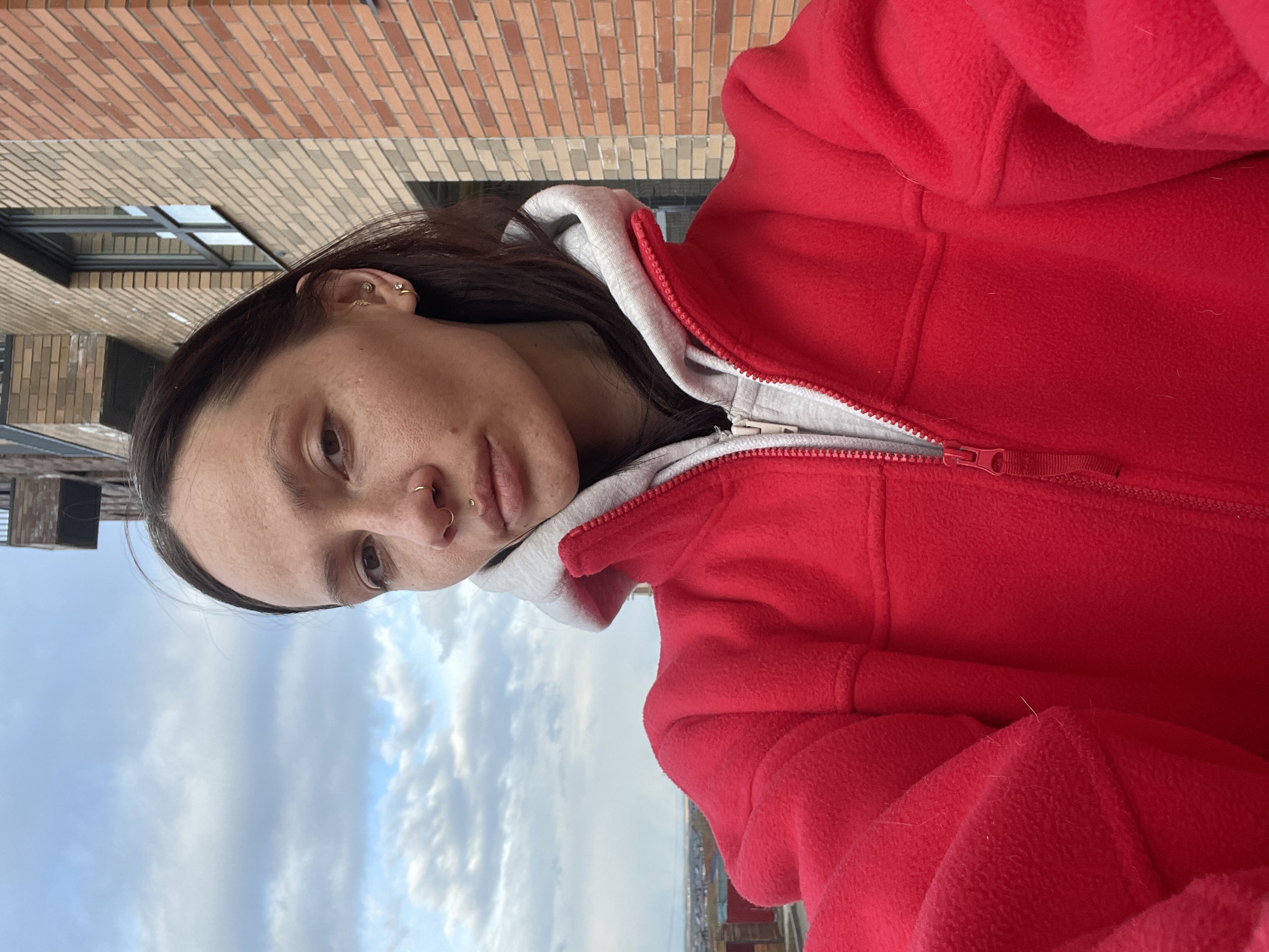 This eye-level, vertical shot features a young person, likely in Izhevsk, Russia, looking directly at the camera. The person has dark hair and multiple facial piercings, including several small hoops and studs in their ears, and a hoop and a stud in their nose. They are wearing a textured red fleece jacket, partially unzipped, revealing a light grey hooded sweatshirt underneath. The person's expression is neutral or contemplative.
The background consists of a brick building with varying shades of brown and beige bricks, including a dark-framed window, occupying the upper portion of the frame. Below this, a bright blue sky with scattered white clouds is visible, suggesting an outdoor setting at a high vantage point, possibly a balcony or rooftop. A blurry cityscape or landscape can be seen in the far distance, just above the horizon line under the clouds. The natural lighting indicates it is daytime. There is no visible text in the image.