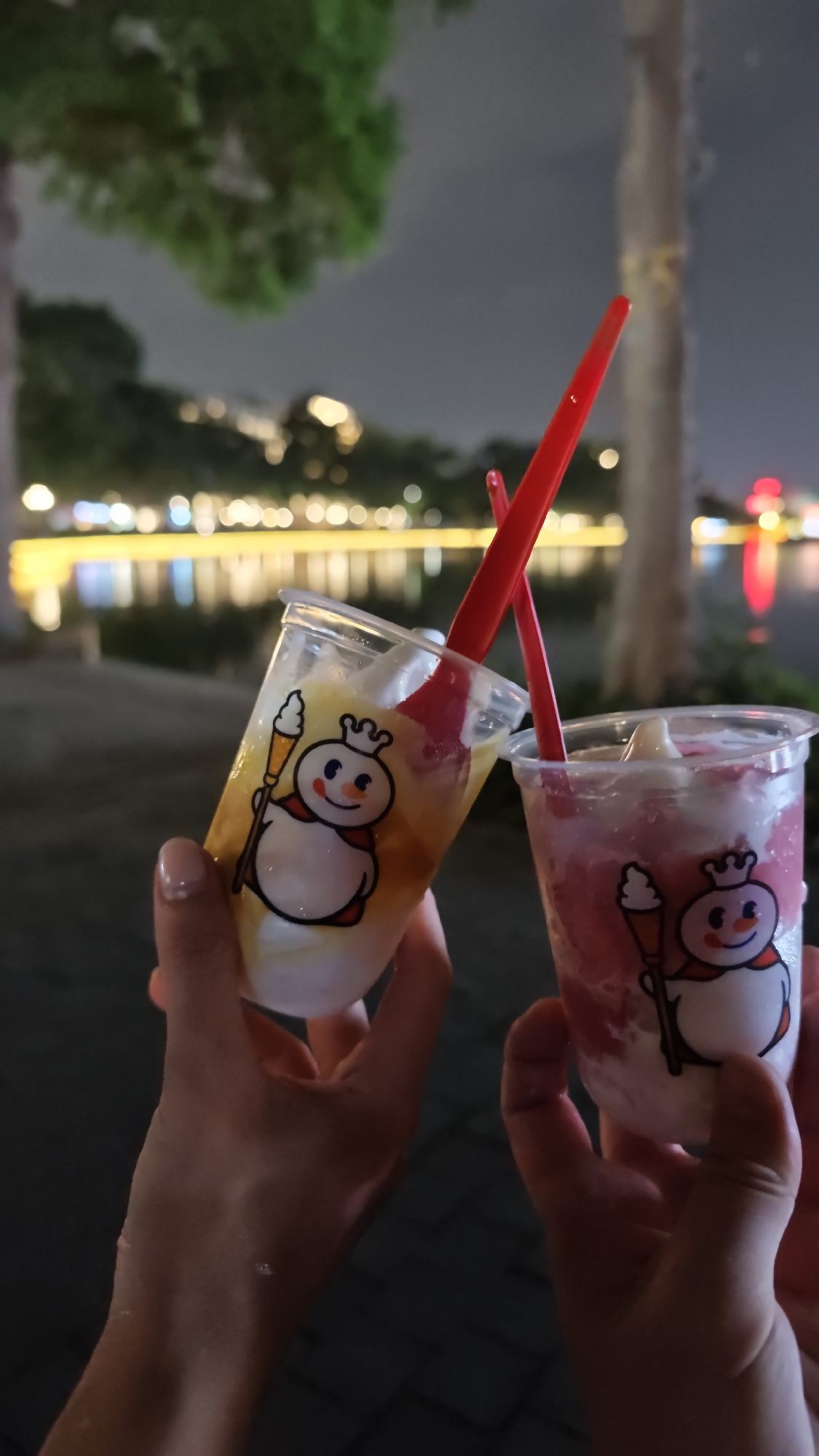Two people hold up two plastic cups of shaved ice with red plastic spoons. The cups have a cartoon logo of a snowman holding an ice cream cone. One cup contains yellow shaved ice and the other cup contains pink shaved ice. The background includes a blurred view of a body of water with lights lining the shore, trees, and an overcast night sky. The location is likely in the city of Hà Nội, Vietnam.