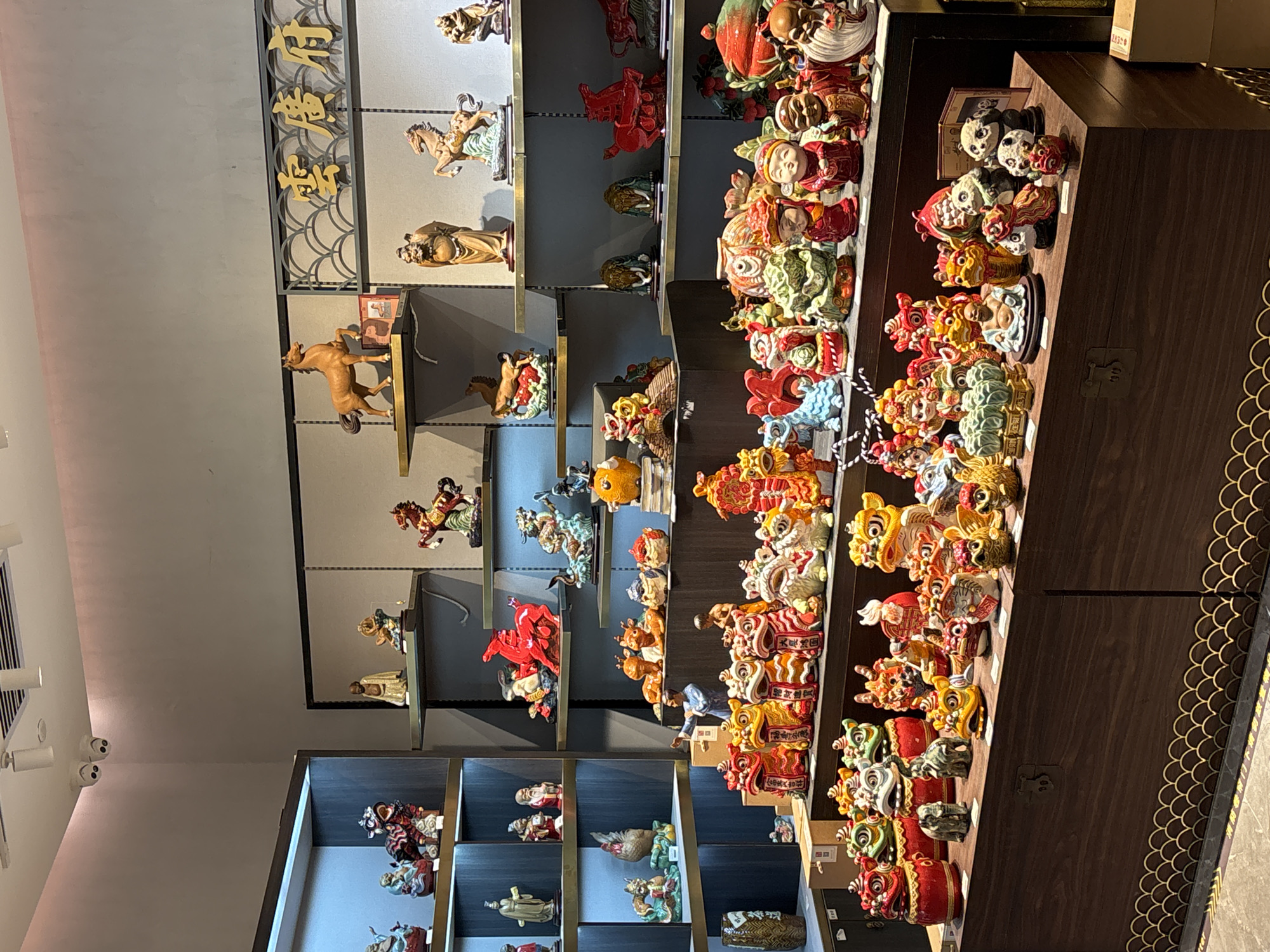 The image shows a store display, likely in Khimki, Russia, featuring shelves stocked with Chinese-themed ceramic figurines. The figurines are colorful and depict various subjects including horses, figures in traditional clothing, and lion dance masks. The shelving units are modern and black, contrasting with the vibrant colors of the figurines. One display case is made of dark wood with a decorative scalloped edge. The store appears well-lit and organized. There are no people visible in the image.