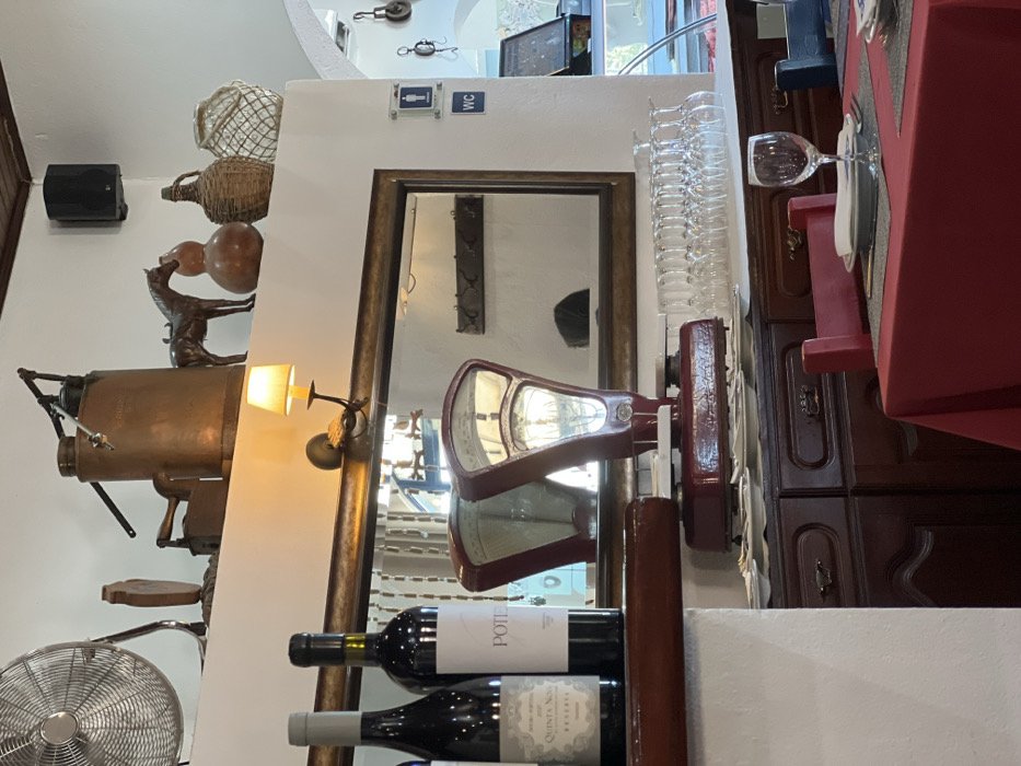 An interior view of an establishment in Coruche, Portugal, displays a dining area and a decorated counter space. In the foreground to the right, a table is set with a red tablecloth and a single place setting: a grey placemat, white plate, folded napkin, fork, knife, and an empty stemmed wine glass. A red wooden-framed chair is partially visible.
Behind the table, a dark wooden cabinet holds a stack of white plates and a prominent red vintage mechanical weighing scale with two dials. To its right, approximately twenty empty stemmed wine glasses are arranged. Above the cabinet, a large rectangular mirror with a dark wooden frame is mounted on a white wall, reflecting a dark coat rack and a blurred form resembling a person's head. A wall-mounted sconce with a yellow-lit lampshade is affixed above the mirror.
To the right of the mirror, two blue square signs on the white wall indicate facilities: an upper sign with male and female pictograms, and a lower one marked "WC." Further right, an arched doorway reveals an area with a display case and hanging objects.
On a wooden ledge in front of the mirror, two dark glass wine bottles are displayed, one partially labeled "QUINTA NOVA RESERVE" and the other "POTE." Above, a white shelf showcases various decorative objects: a large silver industrial fan, a copper manual pump, a dark vintage flat iron, a wooden decorative piece, a dark horse figurine, two gourd-shaped ceramic vessels, and two woven straw-covered bottles. In the upper right corner, a black speaker is mounted, and dark wooden ceiling beams are visible. The scene is brightly lit, with a reflection suggesting a person's presence elsewhere in the room.