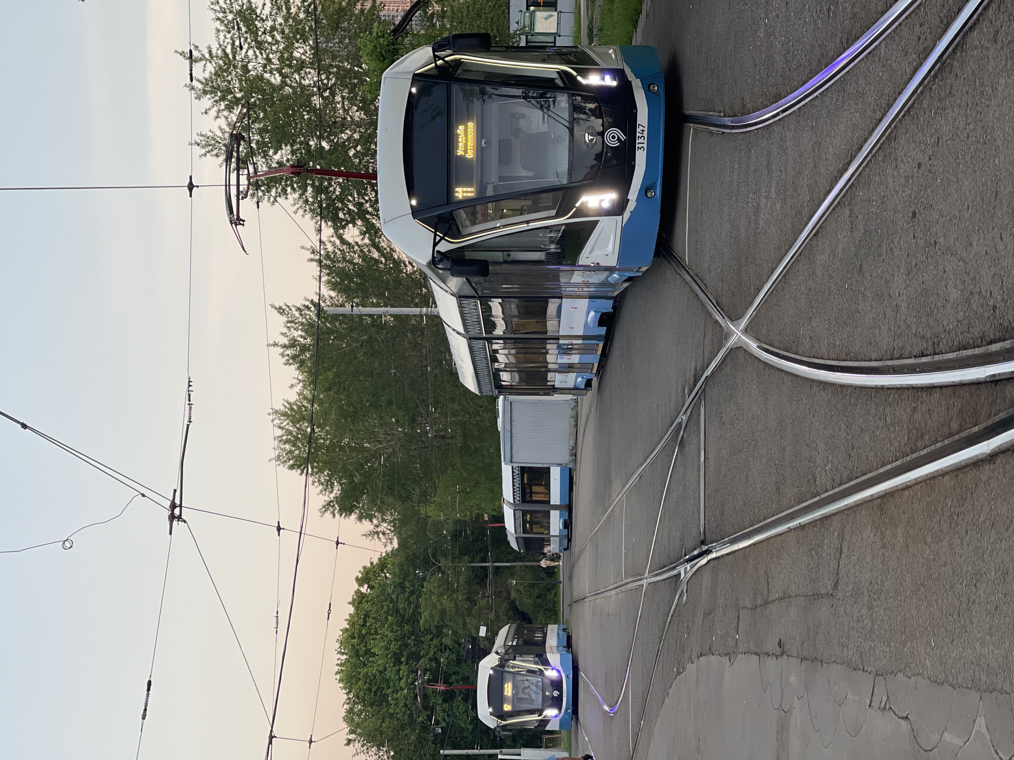 A close-up, angled view depicts a series of white and blue modern trams on a paved track intersection in Unknown, Unknown. The lead tram shows the destination "Усадьба Останкино" on its front display. Overhead power lines extend above the trams, and green trees line the background, suggesting a suburban or park-like setting. The image is oriented vertically.