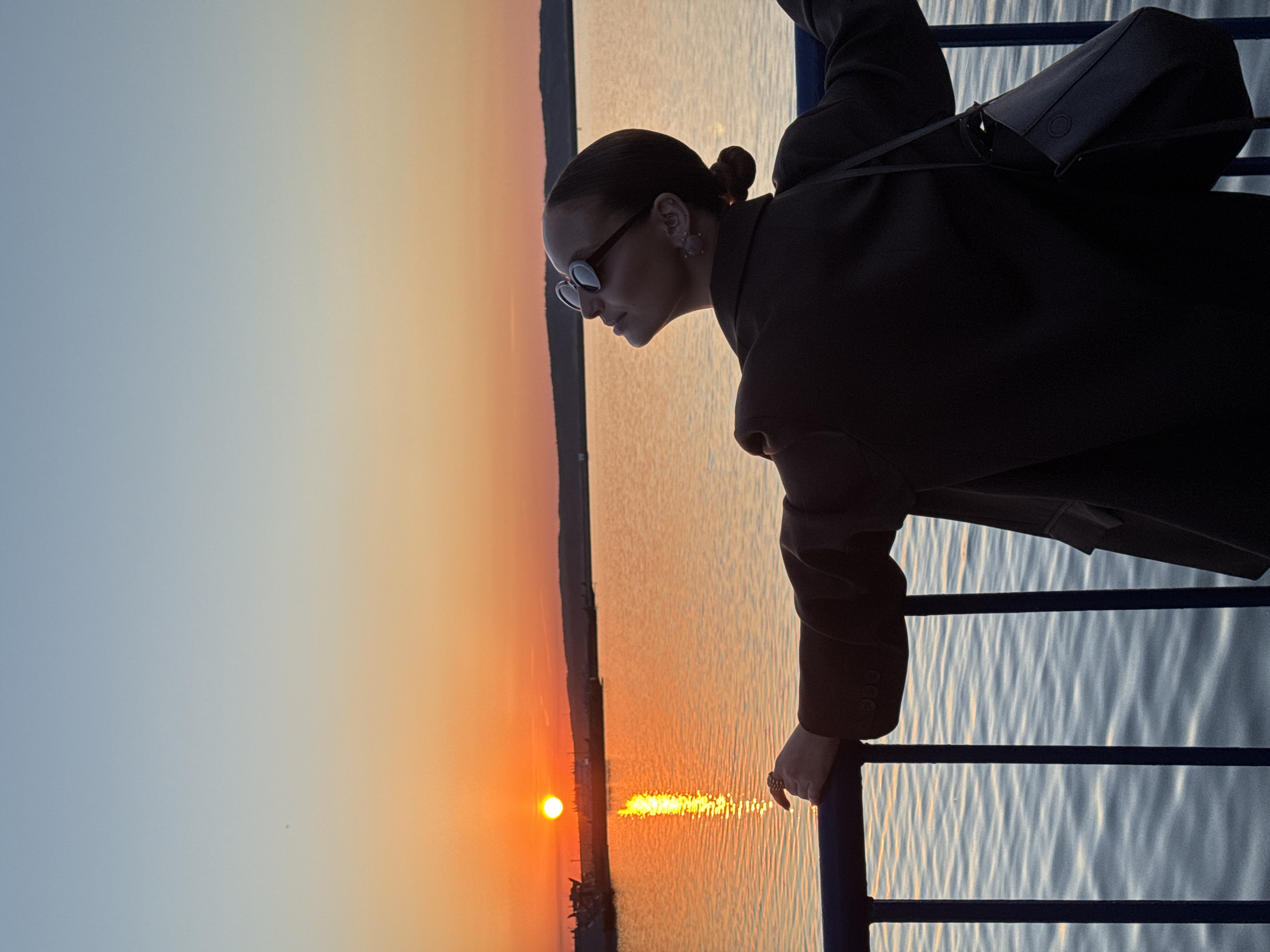 A woman stands on what appears to be a dock or ship railing overlooking a body of water during sunset, possibly in Chaikovsky, Russia. The sun is setting on the horizon, casting an orange and yellow glow on the sky and reflecting on the water. The woman is wearing sunglasses and a dark coat with a bag on her shoulder. She is looking toward the water.