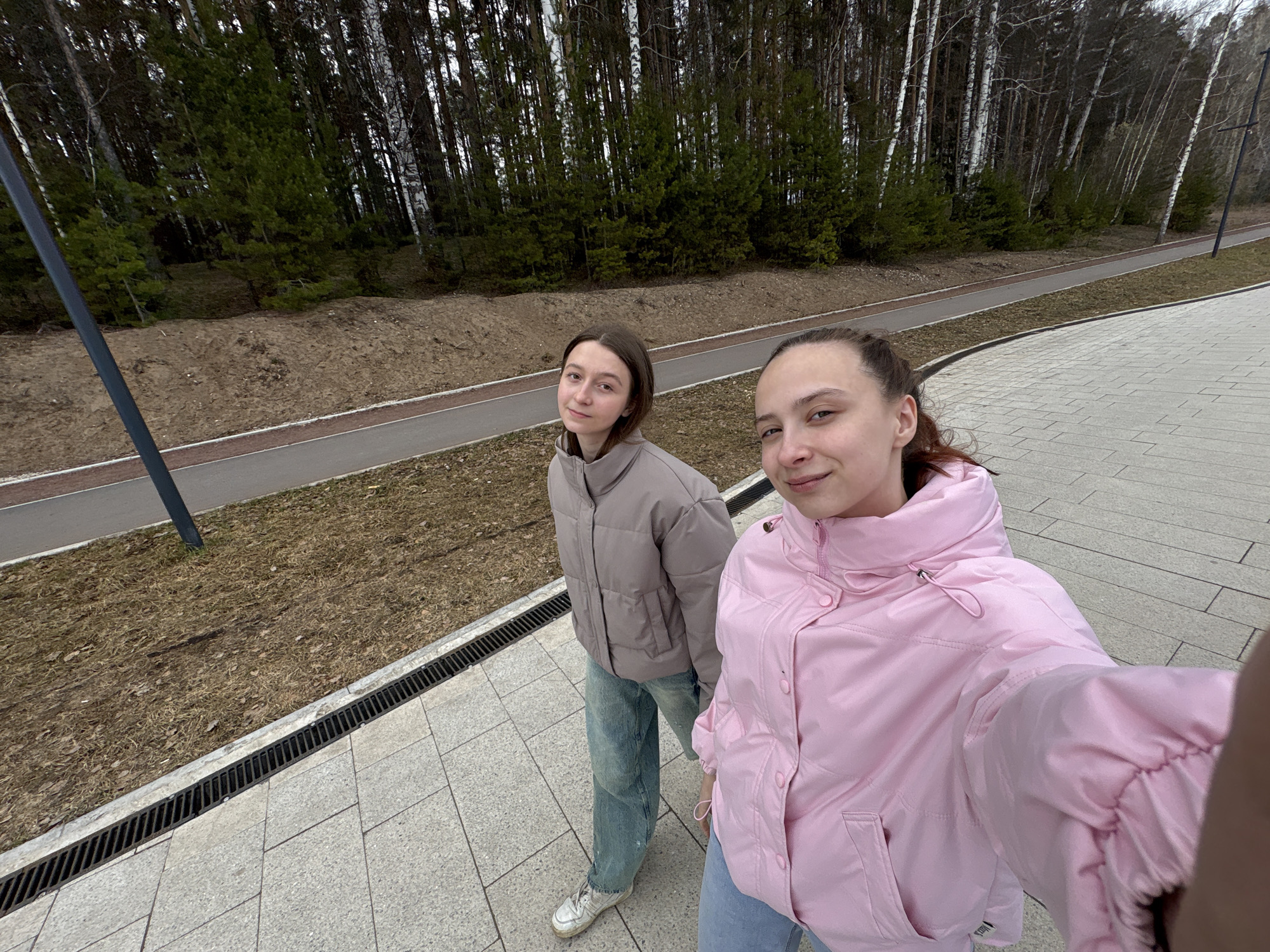 Two young women are standing on a paved area, likely a sidewalk or plaza. The woman on the right is holding the camera, indicating a selfie. She is wearing a pink puffy jacket and jeans. The woman on the left is wearing a similar style jacket in a taupe color, and jeans. Behind them, there is a landscaped area with trees and what appears to be a paved path. The image suggests a location in Chaikovsky, Russia.
