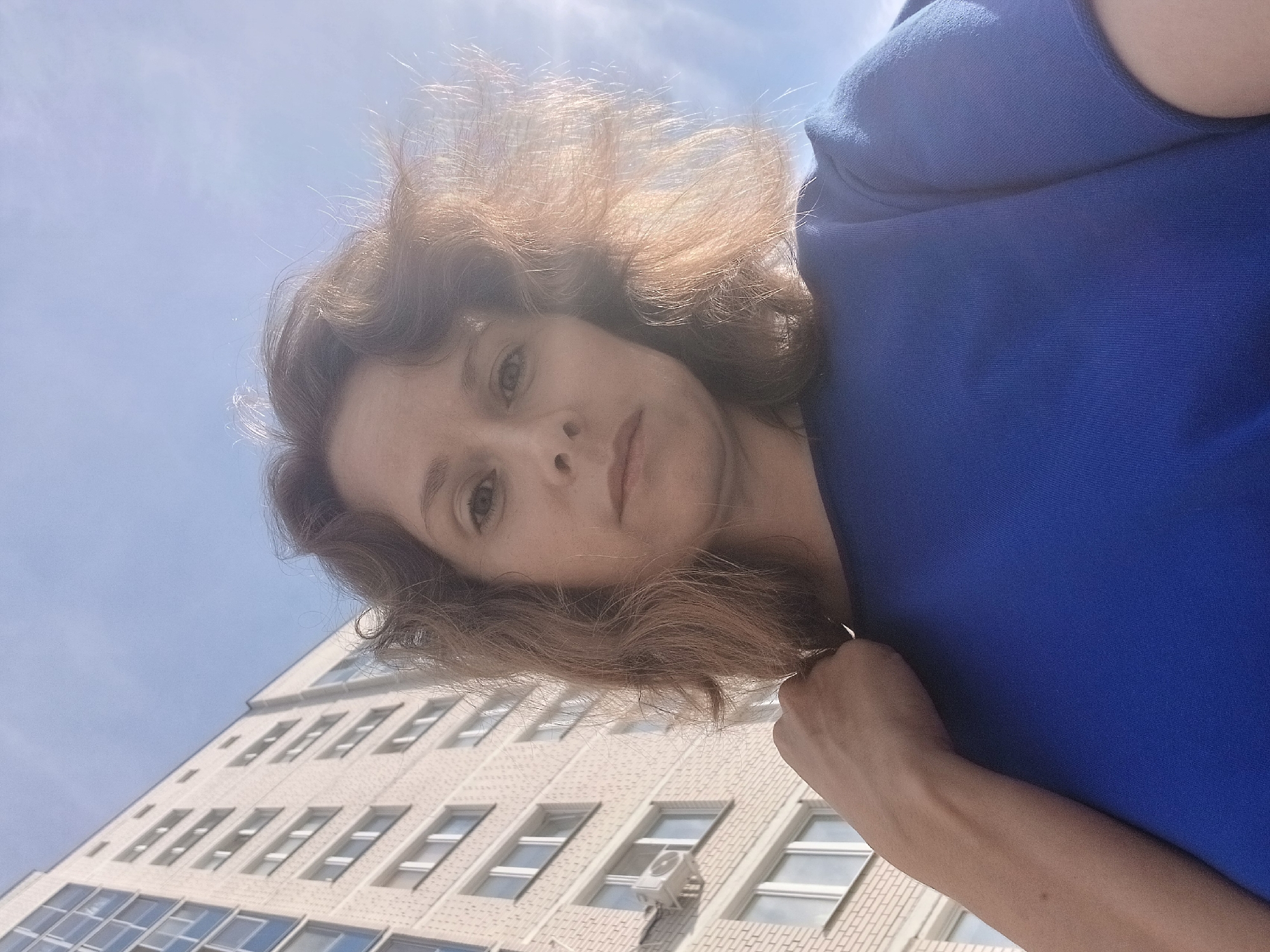 A woman takes a low-angle selfie outdoors with a multi-story building in the background. The building is light-colored with many windows and appears to be an apartment complex. The sky is visible and bright, suggesting it is daytime. The woman has medium-length, wavy brown hair and is wearing a blue t-shirt. Her left hand is raised and touching her hair. The image does not contain enough information to determine the specific location in Yekaterinburg, Russia.