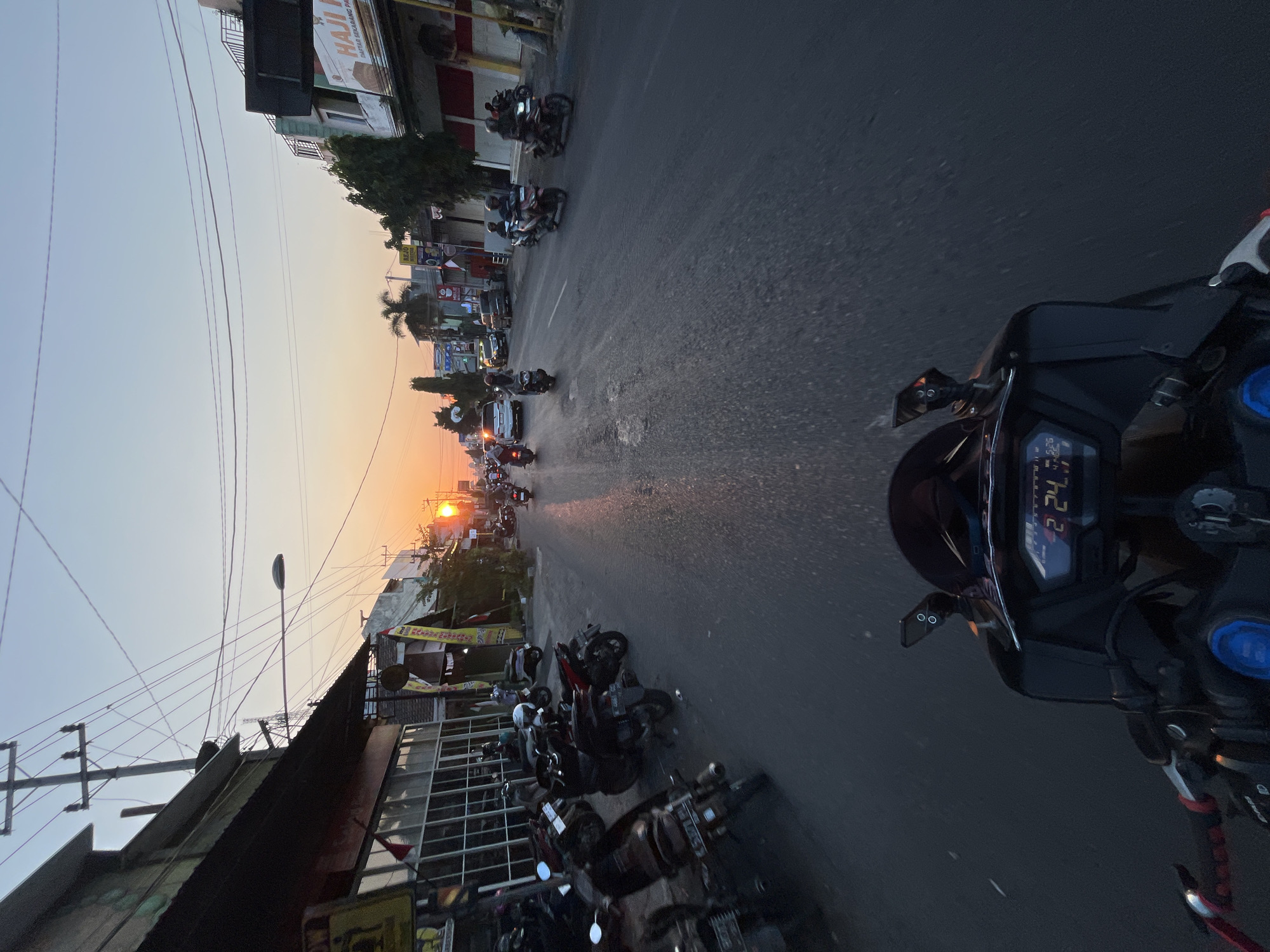 The image depicts a street scene in Welahan, Indonesia, at sunset. The perspective is from the rider of a motorcycle, showing the handlebars and dashboard with a digital display reading "2:24.47". The street is lined with buildings, some appearing to be shops with storefronts and awnings. Numerous parked motorcycles are visible along the left side of the road, and several motorbikes and a white car are in motion on the street. Power lines crisscross overhead, and a street lamp is visible. The setting sun casts a warm glow on the scene, with a clear sky above. The general atmosphere suggests a typical evening commute or activity in a populated urban area.