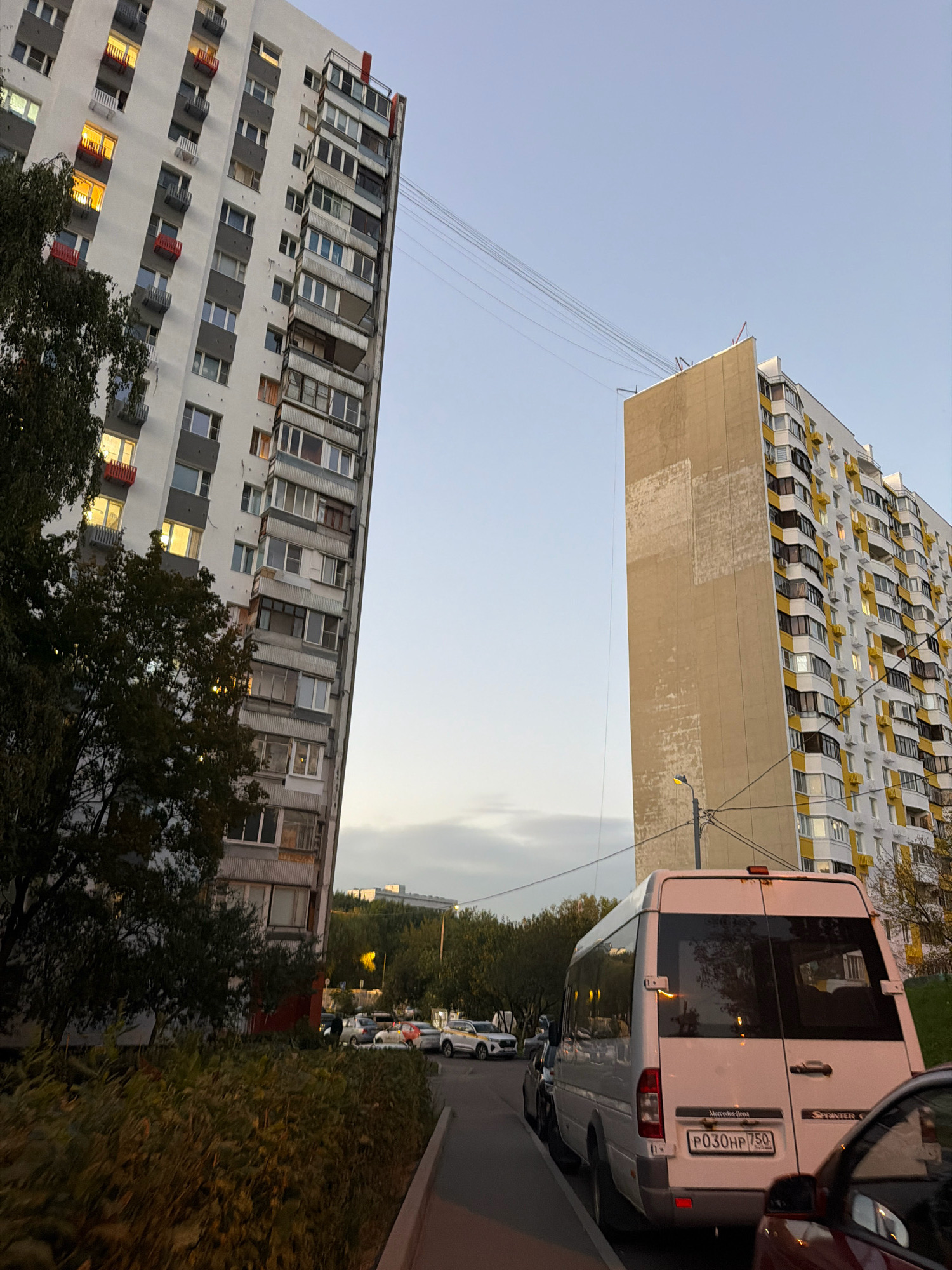 This is an outdoor, eye-level, medium shot image captured in Moscow, Russia, likely during the evening due to the artificial lights visible in the windows of the buildings. The scene depicts a residential area with two tall, multi-story apartment buildings dominating the background. The building on the left is a grey and white structure with numerous balconies, many of which have red or grey railings, and some windows are illuminated with a warm, orange light. A dense, dark green tree is situated in front of this building, partially obscuring it.
To the right, a second apartment building is visible, characterized by a light brown, textured facade on its left side and white and yellow detailing on its right side. This building also features many windows and balconies with yellow railings. Overhead, power lines stretch across the sky between the buildings.
In the foreground, a white Mercedes-Benz Sprinter van is parked on the right side of a paved area, possibly a street or parking lot. The van has a visible license plate that reads "P030HP 750". A dark red car is partially visible in the lower right corner of the frame, parked next to the van. Beyond the van, several other vehicles are parked in a row, including a white SUV. The ground is covered with asphalt, and a sidewalk or curb is present on the left side of the image. Green foliage and trees line the area behind the parked cars, and the sky is a pale blue with some soft clouds visible. The overall impression is a typical urban residential landscape at dusk.