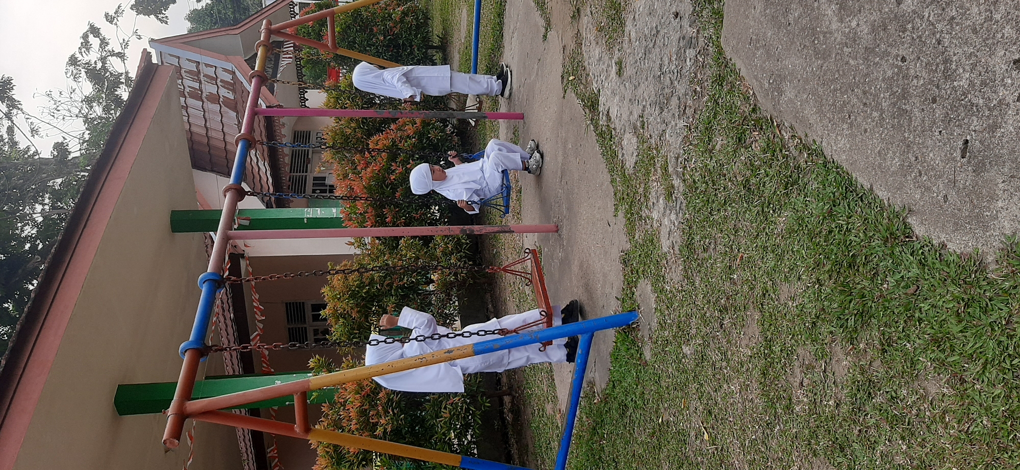 The image depicts an outdoor playground scene with three children wearing white uniforms and hijabs. Two children are on swings, one sitting and the other standing and holding onto the swing chains. The third child is standing beside a swing. In the background, there is a building with a tiled roof and a covered walkway supported by green and pink pillars. Lush green bushes with reddish-orange leaves surround the playground area. The ground is a mixture of concrete and grass. The scene appears to be located in a school or community area within the city of Jambi, Indonesia, suggested by the presence of a building architecture and foliage commonly found in the region.