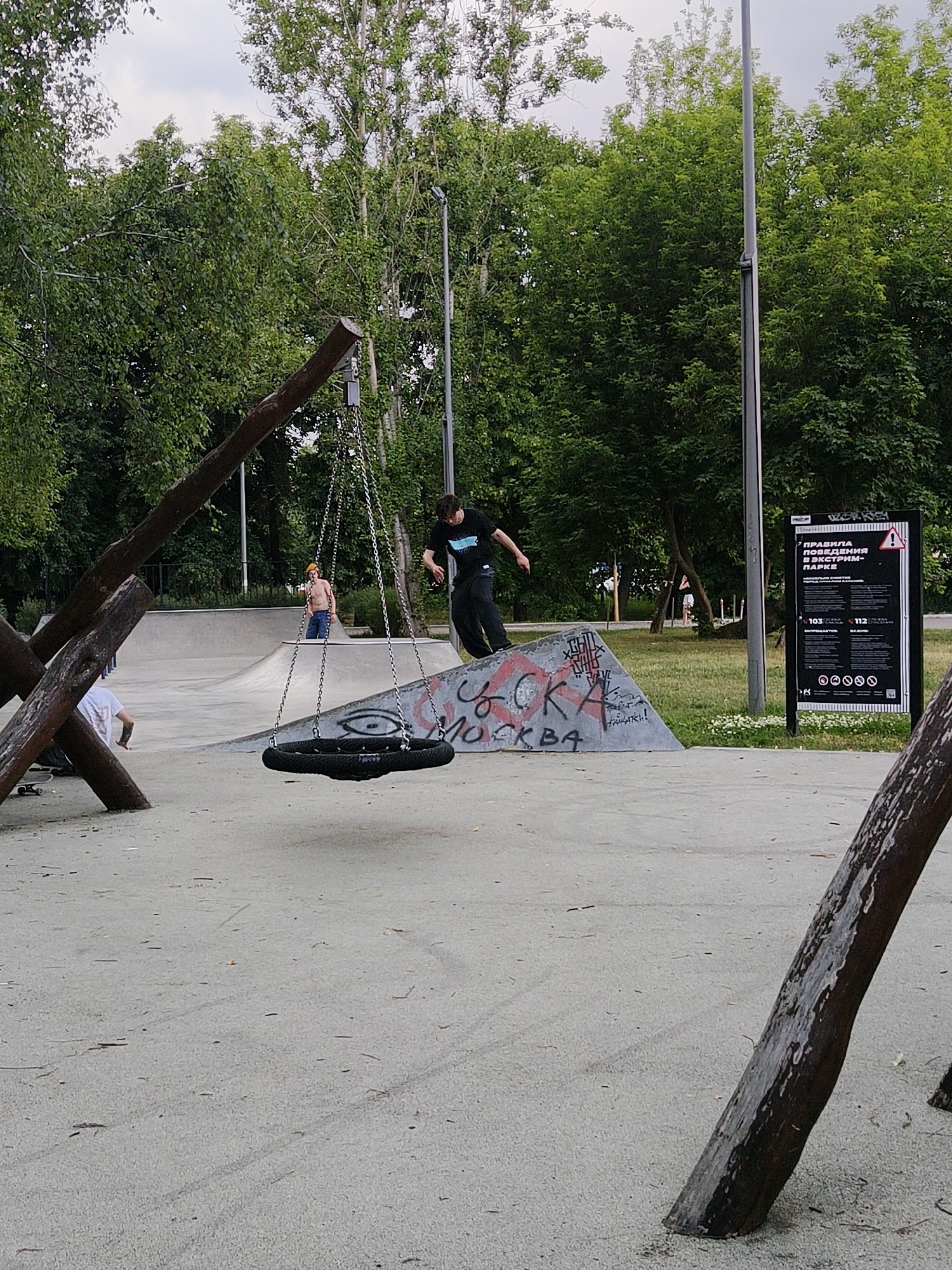 In an outdoor skate park, a person dressed in black is captured mid-air, performing a trick on a ramp. The ramp is adorned with graffiti, including the word "MOSKVA" in red and black spray paint. In the foreground, a tire swing hangs from wooden beams. Behind the ramp, another individual with a bare chest and orange headwear is standing. In the background, a sign with Cyrillic text and a warning symbol is posted. The skate park is surrounded by lush green trees, and the ground is a sandy, light-colored surface. The sky above is overcast.