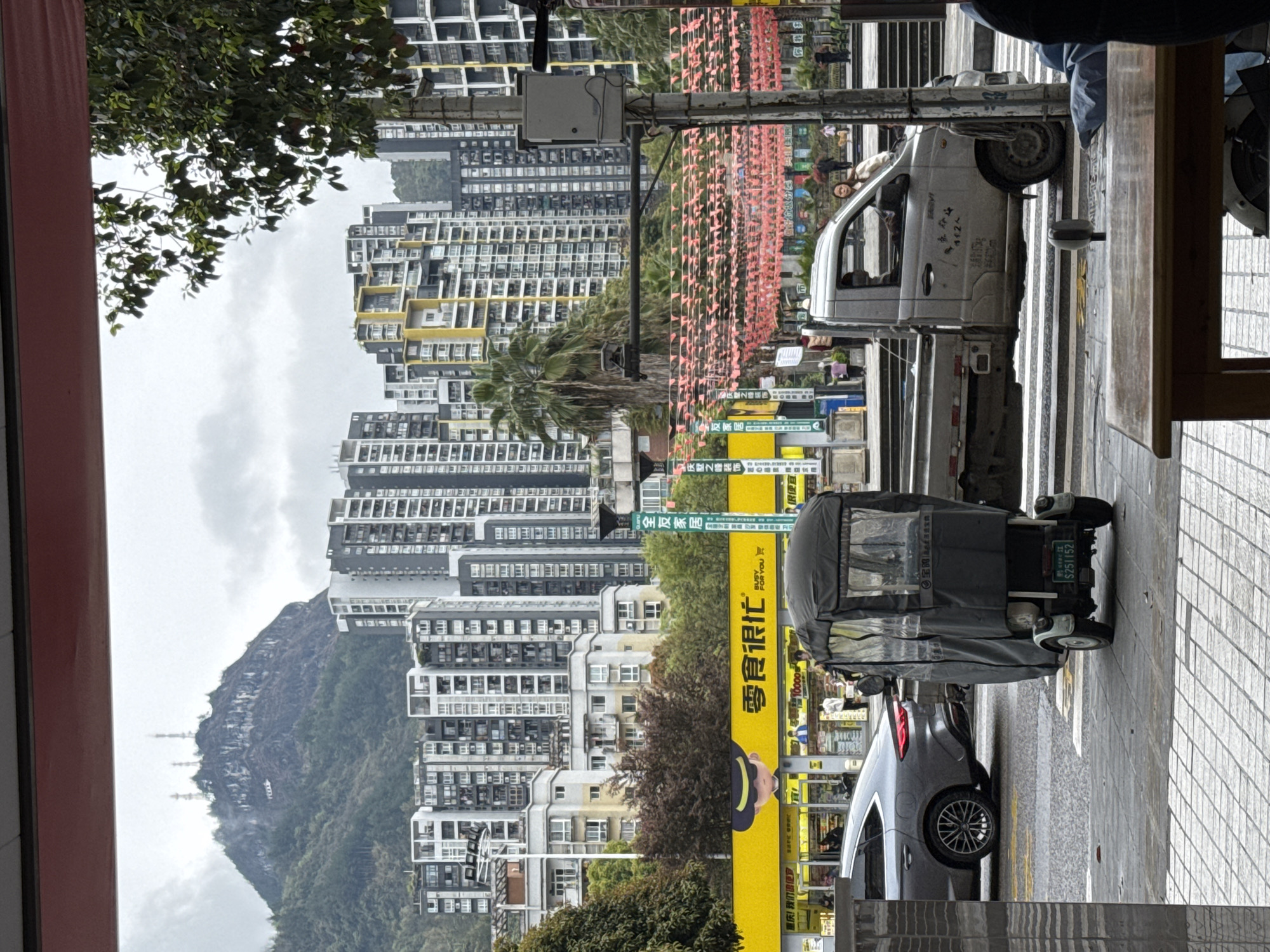 Here's a detailed description of the image:
The image depicts a street scene in what appears to be a densely populated urban area in Qianjiang, China, based on the license plate shown. In the background, there is a mountain partially obscured by clouds and mist, indicating the city's topography. In front of the mountain, a cluster of high-rise residential buildings with a mix of white, gray, and yellow facades dominate the view.
A street runs through the center of the frame. Along the sidewalk, a yellow sign with Chinese characters is visible. A three-wheeled covered vehicle is parked on the sidewalk along with a dark gray car. The sign has a cartoon figure on it and mentions a store called "Zero Snack". A small truck is also visible in the scene. A string of orange triangular flags is strung above the street, likely for a celebration or decoration. There's a street sign on the street pole.
The image is shot from an elevated point of view, possibly from a building or an upper floor of a shop. The day seems overcast and possibly humid. The sidewalk is paved with gray tiles.
