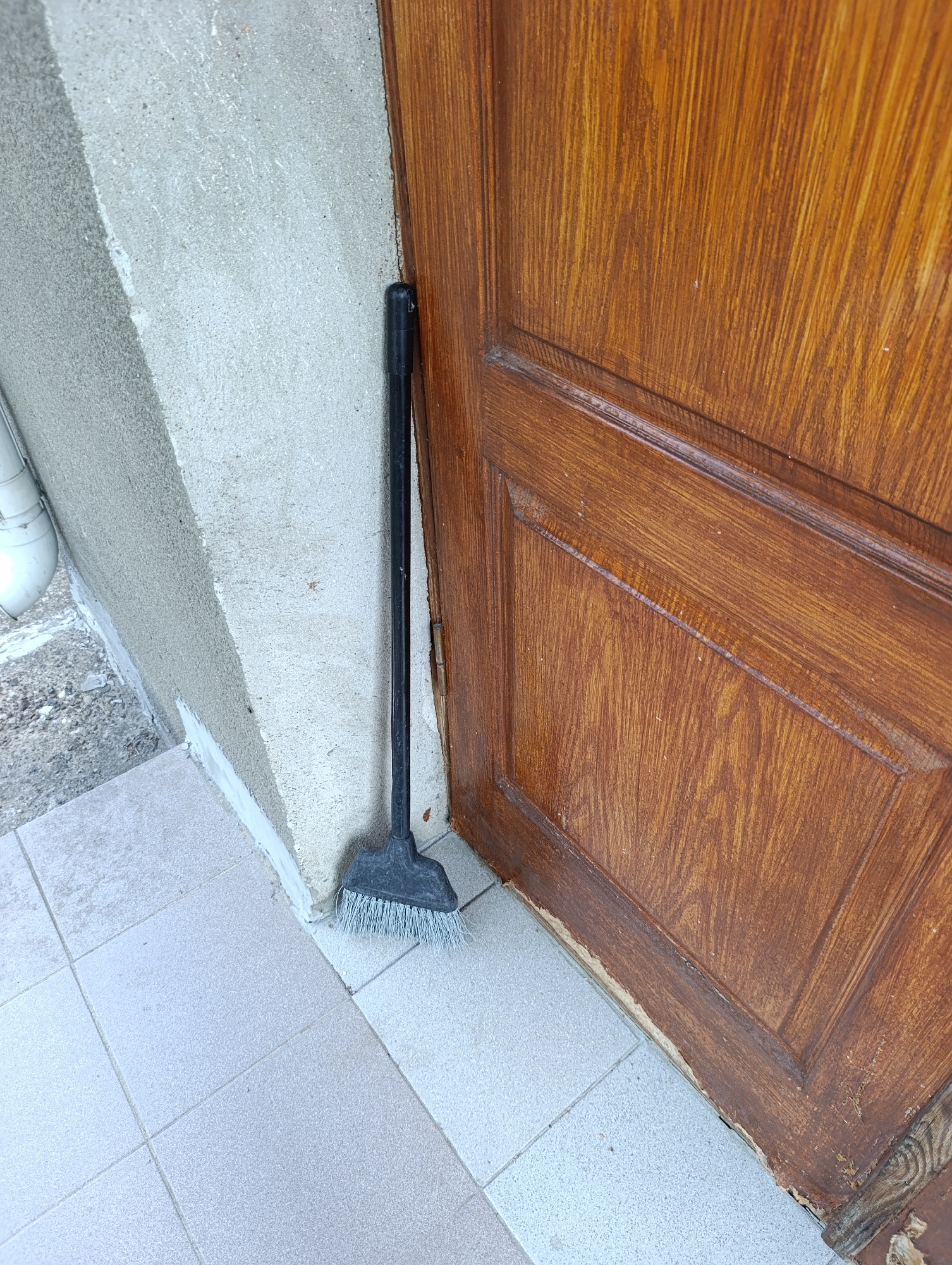A black broom with a wide, grey plastic head and white bristles leans against a textured, light-colored concrete wall. To the right of the broom is a dark brown wooden door with a visible grain and paneling. The broom's handle is angled upwards, resting against the wall. The floor in front of the wall and door is tiled with light grey square tiles, some appearing slightly worn. The scene is outdoors, suggesting an exterior entrance. The provided coordinates place this scene within the Dubăsari town council, Moldova. There are no people or animals visible in the image.