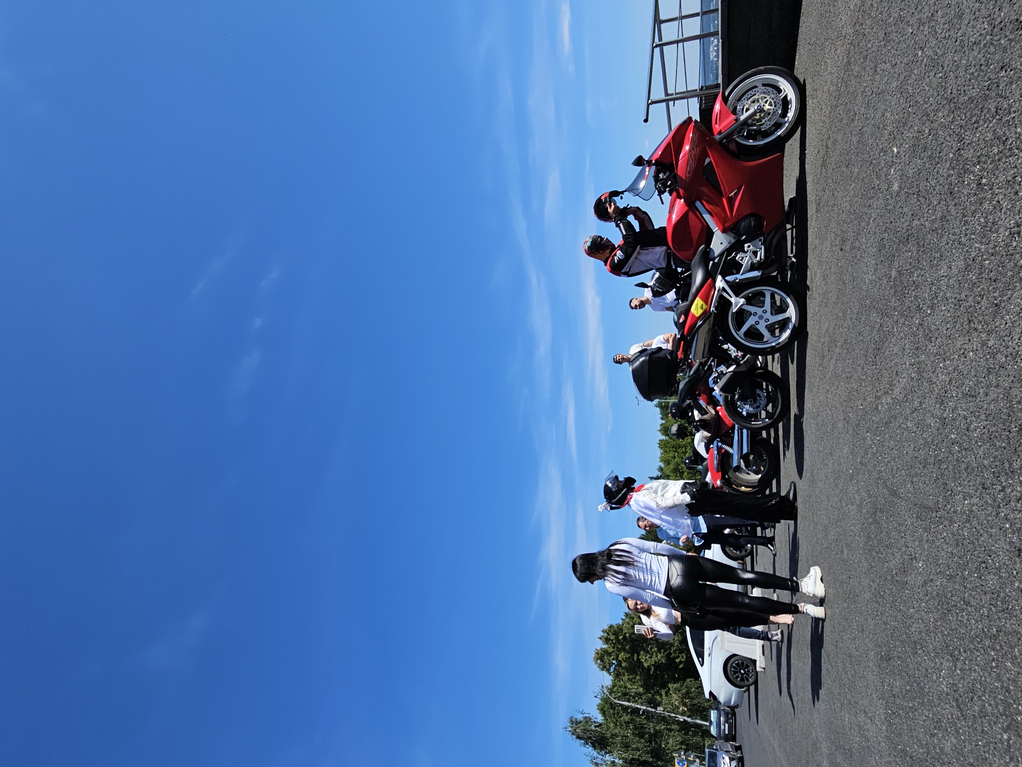 On a bright, clear day with a vibrant blue sky, a group of people gathers around several motorcycles parked on an asphalt surface, possibly a waterfront promenade or parking lot in Ulyanovsk, Russia. The scene appears to be outdoors, with trees and foliage visible in the background on the left, and a metal railing overlooking a body of water on the right.
In the foreground, a striking red motorcycle is prominently displayed, with a person in a racing jacket and helmet adjusting the helmet. Beside it, another motorcycle, partially obscured, is also visible. To the left, a woman with long dark hair, wearing a white long-sleeved top and tight black pants, is posing for a photo being taken by another woman. A man in a white shirt and black pants stands nearby. Further to the left, a white car is parked.
The overall atmosphere suggests a gathering or meeting, possibly related to motorcycles, given the presence of multiple bikes and people dressed in riding gear. The clear sky and ample sunlight indicate pleasant weather conditions. The perspective of the shot is low, emphasizing the motorcycles and the people around them against the expansive sky.