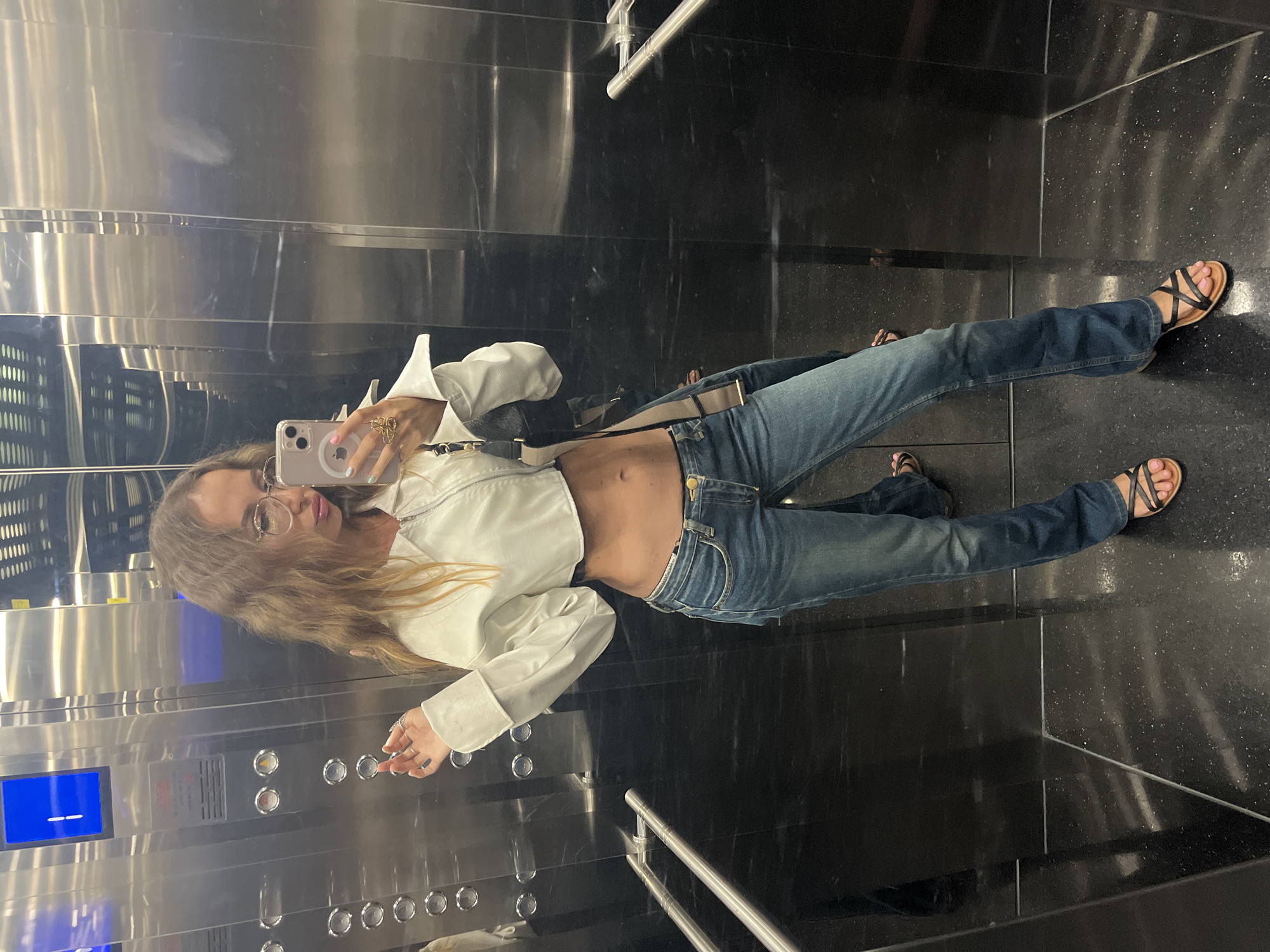 A young woman is seen taking a mirror selfie inside a modern elevator, likely in Đà Nẵng, Vietnam. She has long, light brown hair and wears round glasses, with light pink lipstick and pastel-colored nail polish (pink, light blue, light green) on her fingernails. Her outfit consists of a white cropped jacket that exposes her midriff, blue jeans, and black sandals with light pink painted toenails. Rings are visible on her fingers.
She holds an iPhone with a clear case in her right hand, pointing it towards the reflective surface to capture her image. A shoulder bag with a visible strap and gold buckle hangs from her shoulder. The elevator's interior features sleek, dark, highly reflective walls, possibly polished stone or metal, interspersed with brushed metallic panels. The floor is made of a dark, speckled material, indicative of granite or similar stone. A metallic handrail extends along the right side of the elevator.
To the woman's left, the elevator control panel is visible, featuring a series of circular buttons and a blue digital display showing a white horizontal line, though no specific text is clearly readable. The setting appears clean and contemporary, characteristic of a modern building such as a hotel, apartment complex, or shopping center. The overall impression is a casual, everyday moment captured in an urban transitional space.