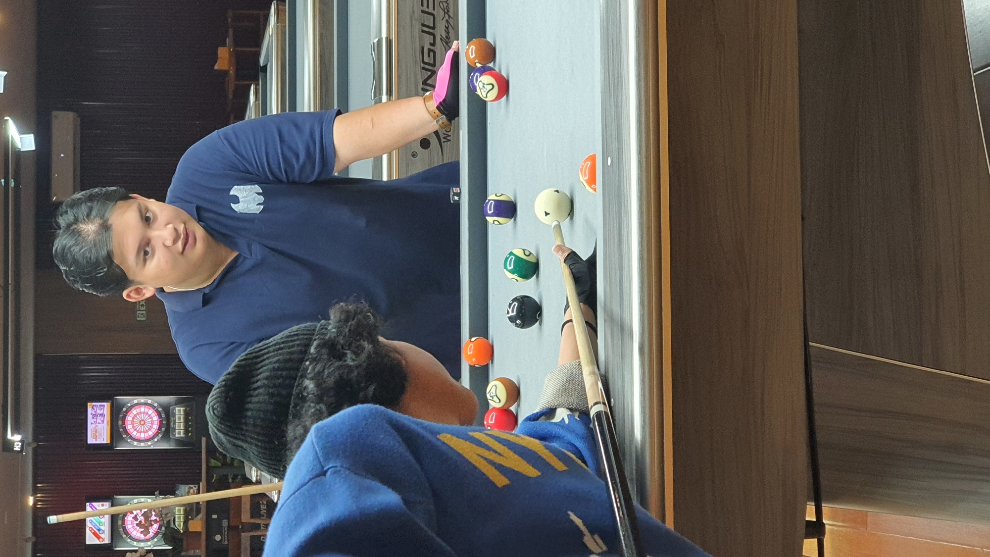 Two individuals are positioned at a billiards table in what appears to be an indoor entertainment venue in Kuta Selatan, Indonesia. One person, wearing a blue hoodie and a black beanie, is actively playing, holding a pool cue and aiming at the cue ball. This individual is wearing a black glove on their shooting hand. The other person, wearing a navy blue polo shirt with a white logo and a pink and black wristband on their right arm, is observing the game, looking towards the playing individual. The billiards table is covered in a gray felt surface and is populated with several colored billiard balls. In the background, there are dartboards mounted on the wall, suggesting the venue offers multiple recreational activities. The overall environment suggests a casual, recreational setting.