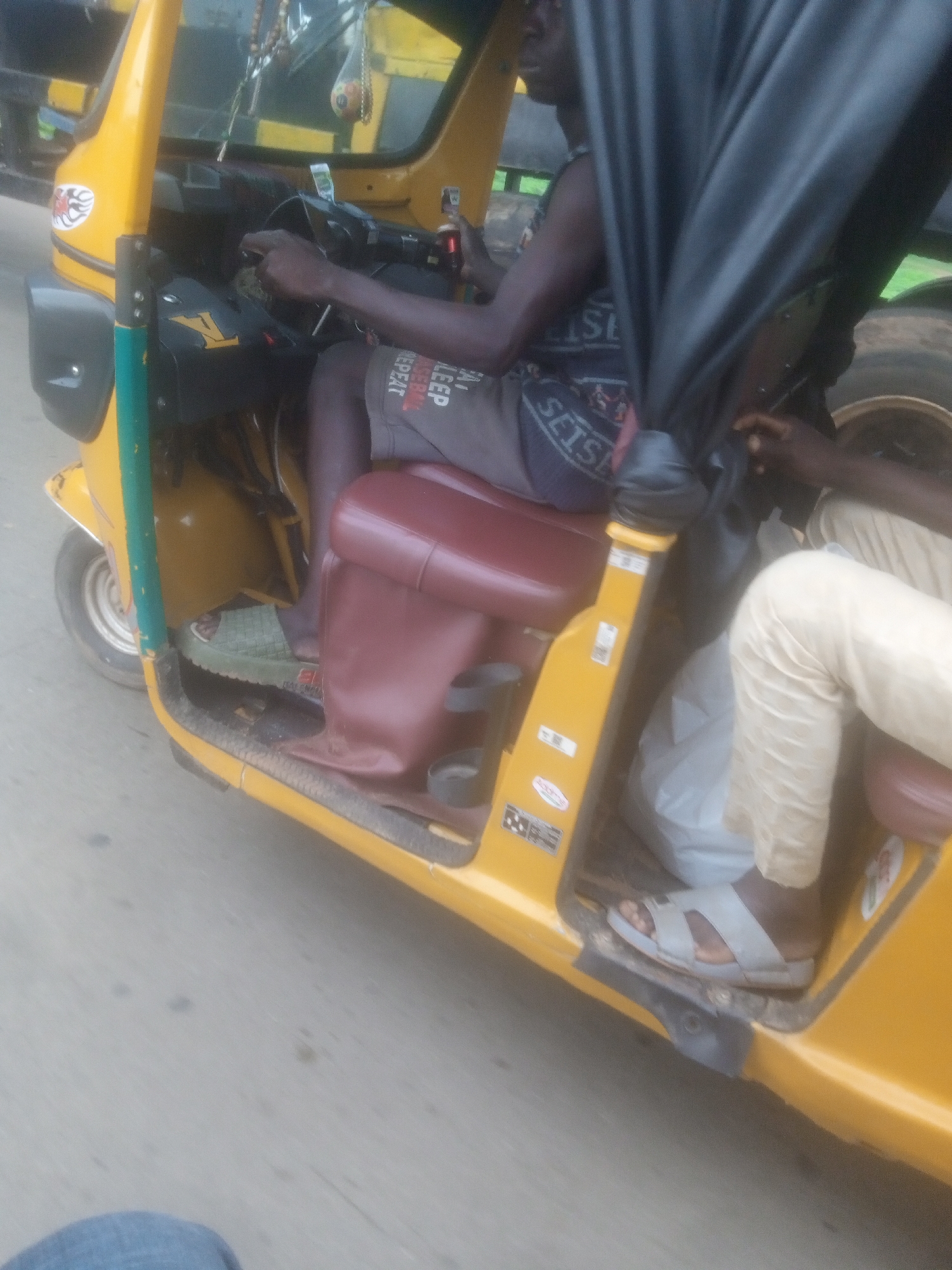 The image depicts the interior of a yellow three-wheeled auto-rickshaw, commonly known as a "Keke Napep," in Nassarawa, Nigeria. A dark-skinned male driver, wearing a dark sleeveless top and grey shorts with text that reads "KEEP REPEAT," is seated at the controls. His hands are on the steering wheel. To the right of the driver, another dark-skinned individual is seated, wearing a light-colored long-sleeved shirt and beige pants. This passenger's legs are visible, with one foot wearing a light-colored sandal. The auto-rickshaw has red cushioned seats. A black fabric canopy is partially obscuring the view. The exterior of the auto-rickshaw is yellow, with some green trim on the front. The vehicle appears to be on a paved road, suggested by the grey, speckled surface visible in the foreground and background. There is a faint green blurred background, possibly indicating vegetation or other vehicles.