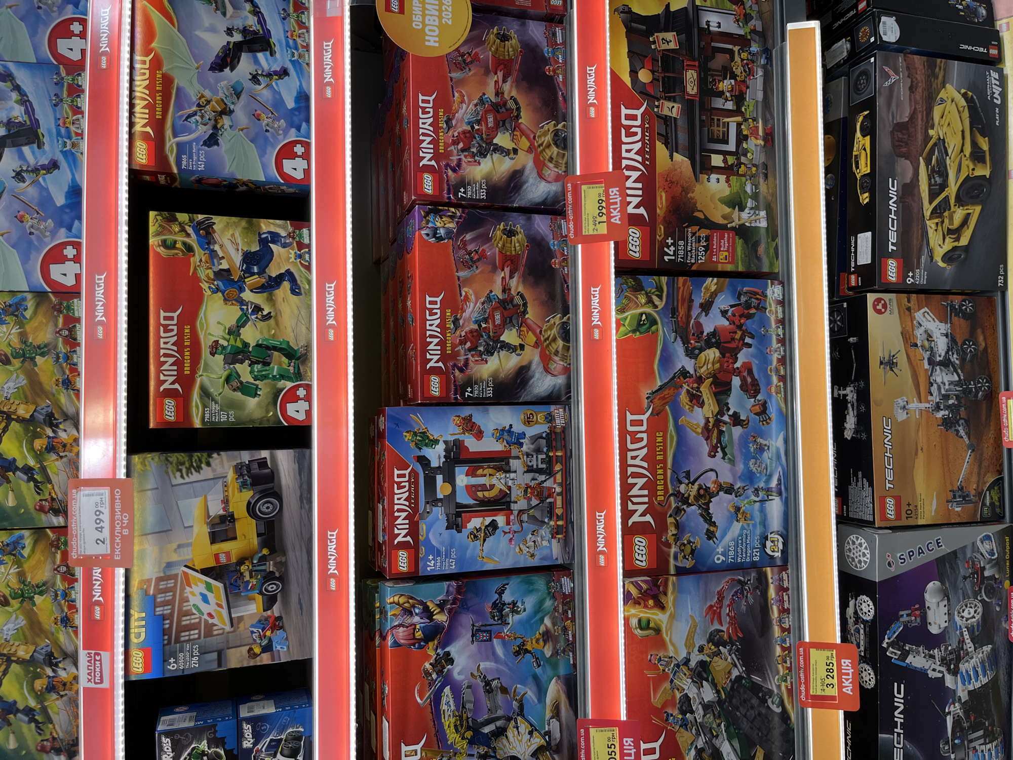 The image shows a retail store shelf displaying various LEGO toy sets. The shelves are illuminated by overhead lighting and a red LED strip along the front edge. The LEGO sets are predominantly from the Ninjago and City lines, with some Technic sets visible at the bottom. The Ninjago sets feature dragons, mechs, and ninja characters, with prominent branding and age recommendations (e.g., "4+", "7+", "14+"). Some boxes have sale stickers or promotional tags. One such tag displays a price of "2 499.00 грн". Another tag shows a price of "3 285.00 грн" with "АКЦІЯ" (meaning "SALE" in Ukrainian). The environment appears to be a typical toy aisle in a large retail store. The presence of Ukrainian currency ("грн") and the Ukrainian word for sale suggests the location is in Ukraine. Given the explicit mention of Kharkiv in the prompt, it is likely this scene is within a store in Kharkiv. No people are clearly visible interacting with the products. The arrangement is orderly, with boxes stacked neatly.