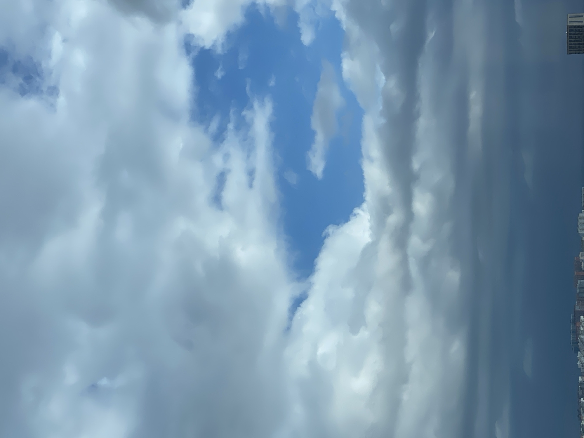 The image captures a cloudy sky over a city, likely Krasnodar, Russia. The sky is predominantly filled with various types of clouds, ranging from fluffy white cumulus formations to darker, more ominous stratus clouds. In the upper portion of the frame, a patch of bright blue sky is visible, suggesting a break in the cloud cover. Below this, the clouds are thicker and appear to be gathering, with some areas exhibiting a greyish hue. The lower third of the image shows a distant cityscape, with buildings of varying heights visible. The overall impression is one of a somewhat overcast day with the potential for changing weather. There are no people or visible actions depicted.