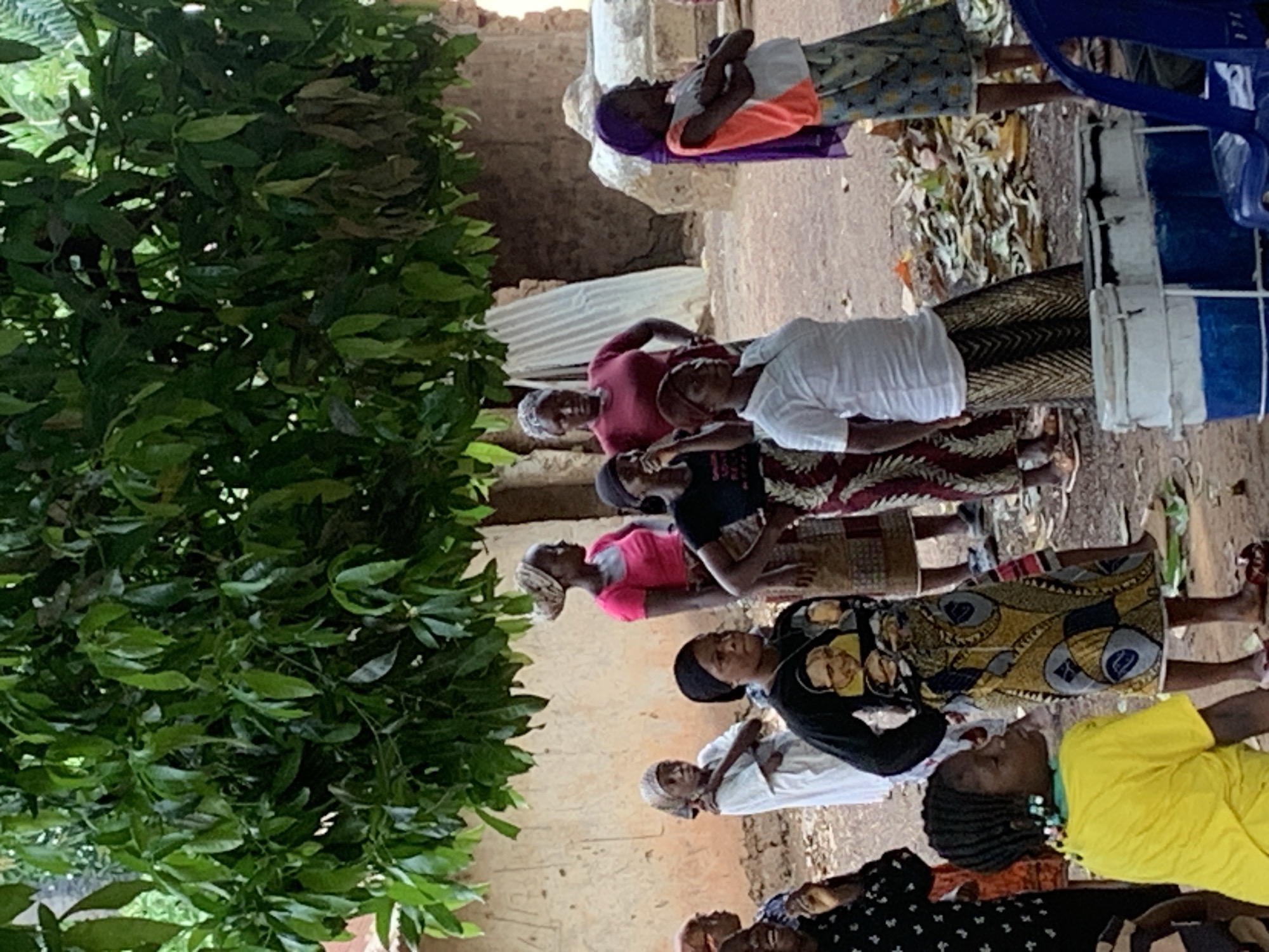 A medium shot depicts a group of African women standing outdoors, likely in the city of Akwanga, Nigeria, given the context. The women are dressed in a mix of traditional and Western clothing. Some wear head coverings. They are standing on a dirt surface with scattered debris. A green bush is to the left. In the background, there's a dilapidated building with a partially collapsed wall. A plastic blue chair and a white bucket are visible on the right. The interaction between the women is unclear from the image. The overall environment suggests a rural or semi-rural setting.