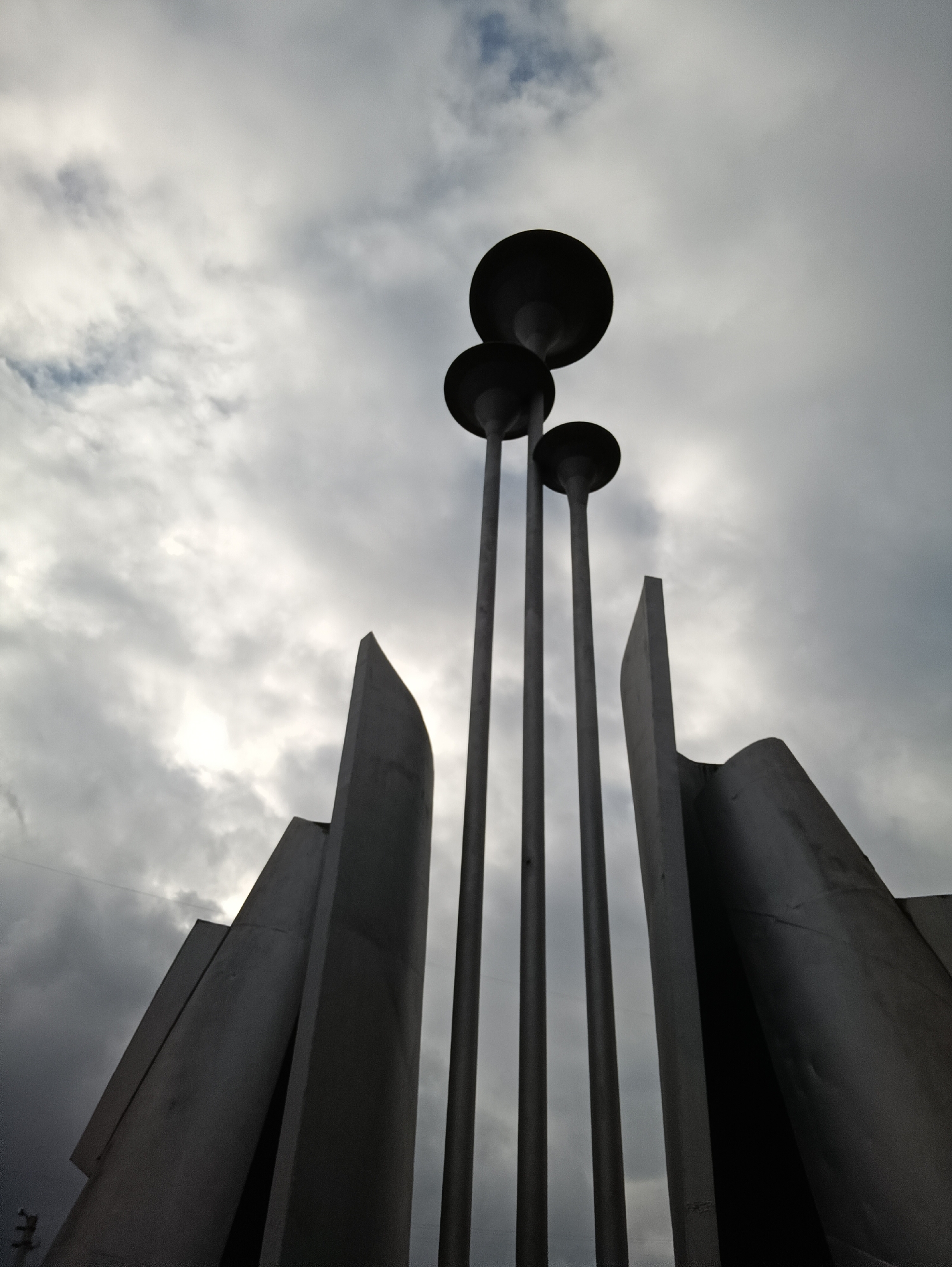 A low-angle shot captures a modern monument or sculptural installation in Chaikovsky, Russia, against an overcast sky. The central feature comprises three tall, slender poles, each topped with a dark, broad, dish-shaped element, likely parts of a lighting system or symbolic design. These poles stand in stark silhouette against the bright, cloudy sky, which displays varying shades of gray, indicating dense cloud cover.
On either side of the poles, large, geometric structures made of concrete or metal rise. These components feature sharp, angular edges and some subtly curved surfaces, contributing to an abstract and monumental form. The structures appear to be silhouetted against the sky, with the light source seemingly behind the clouds, casting a diffused glow.
The scene is devoid of human presence or activity, lending a quiet and solemn atmosphere. The cloudy conditions suggest an overcast day. No text is visible in the image. The overall impression is one of modern architectural design and monumentality.