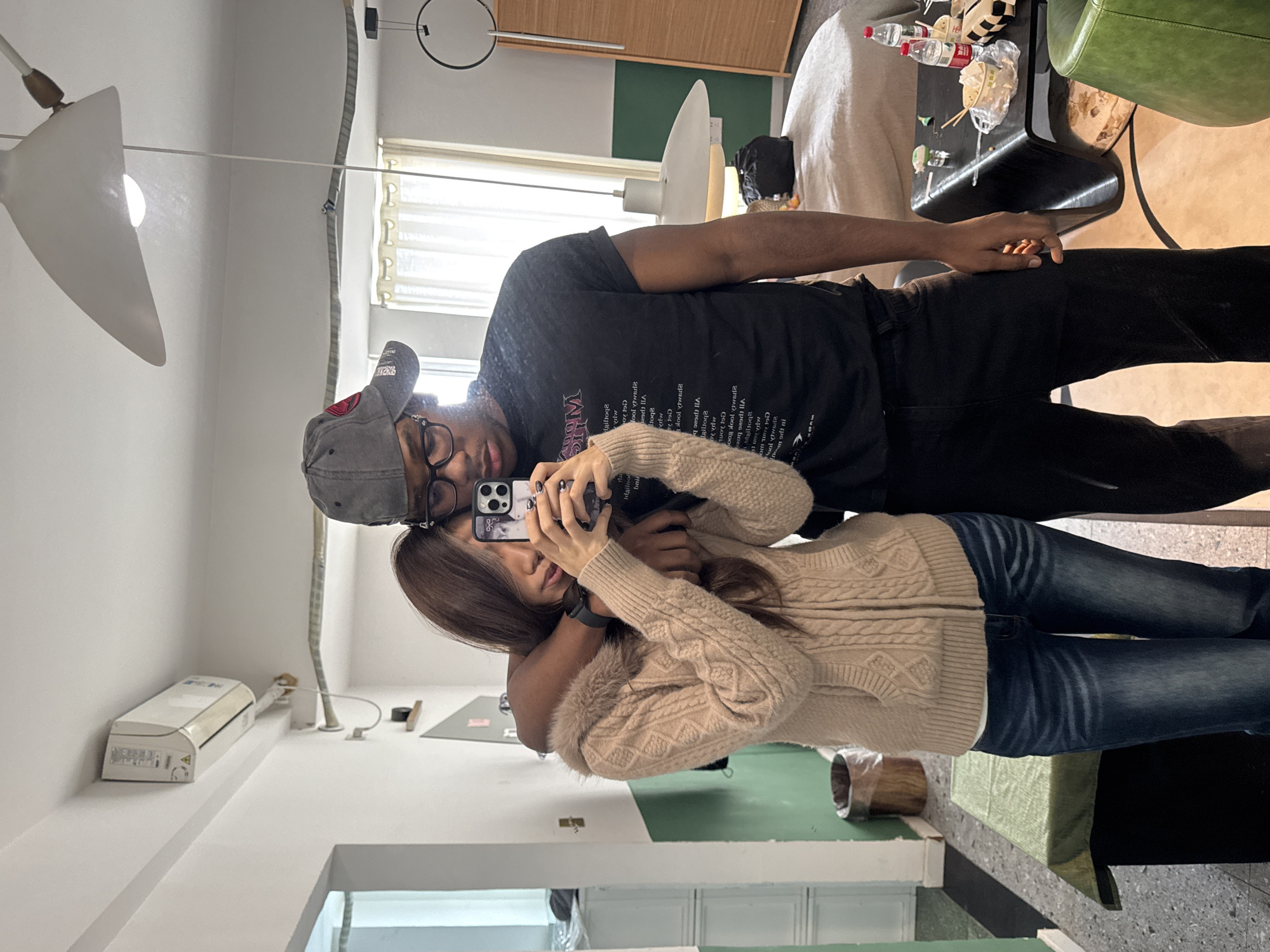 A mirror selfie taken indoors in a modern living space or lounge area, located in Liangjiang, China. Two individuals are present: an East Asian female and a male of African descent.
The female, with long brown hair, wears a light beige cable-knit hooded cardigan with faux fur trim, a white undershirt, and dark blue jeans. Her left hand holds a black smartphone with a prominent camera module, obscuring part of her face. She wears a dark smartwatch on her left wrist, and her fingernails are painted dark. Her right arm is around the male's waist.
The male wears a gray baseball cap with a red emblem, black-rimmed glasses, a black short-sleeved t-shirt with white text (reversed), and dark pants. His right arm is around the female's shoulders.
The room features white walls, with a lower section painted dark green in certain areas. The floor in the foreground is a speckled gray terrazzo or concrete, transitioning to a lighter floor further back. A white wall-mounted split-unit air conditioner is visible on the upper left. A large, white, inverted bowl-shaped pendant light hangs from the ceiling in the mid-right, alongside a minimalist black circular pendant light structure. A silver-colored conduit runs along a wall/ceiling intersection.
Furniture includes a rectangular black table draped with green fabric and a brown cylindrical trash can in the lower left. In the background, there is a large, rounded, low-profile bed or sofa in a light color, with a black bag resting on it. In the foreground right, a distinctive black, low coffee table holds two plastic water bottles, a clear plastic bag containing white takeout containers, chopsticks, and a small black and white checkered item. A portion of a green armchair is visible in the lower right. A window with sheer white curtains allows natural light into the room. A tall, light-colored wooden cabinet stands on the far right.