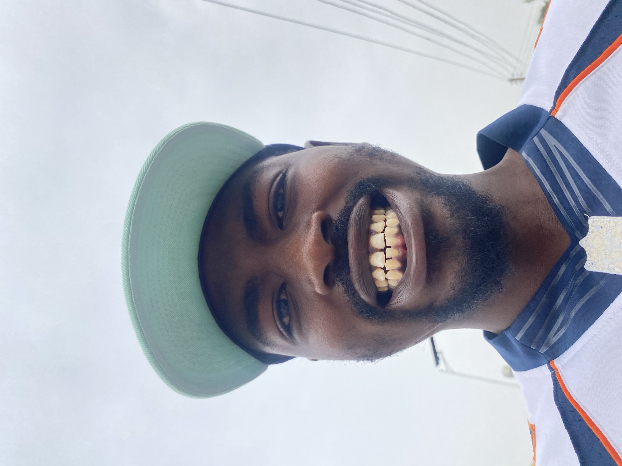 The original image displays a man from a low-angle perspective, smiling brightly and looking directly at the camera. He has a dark complexion and a beard, and is wearing a light green baseball cap. His attire includes a white polo-style shirt with a dark blue collar and blue accents on the shoulders and sleeves, detailed with orange and blue stripes, reminiscent of a sports jersey. His wide, cheerful smile reveals his teeth. The background is a uniformly bright, light grey or white sky, suggesting an outdoor setting on a clear or overcast day. Faint white lines or structures are visible in the upper portion of the background, possibly wires or rigging. The man's expression conveys happiness and a friendly demeanor. There is no visible text on his clothing or cap. The scene is situated in Barakin Akawo, Nigeria.