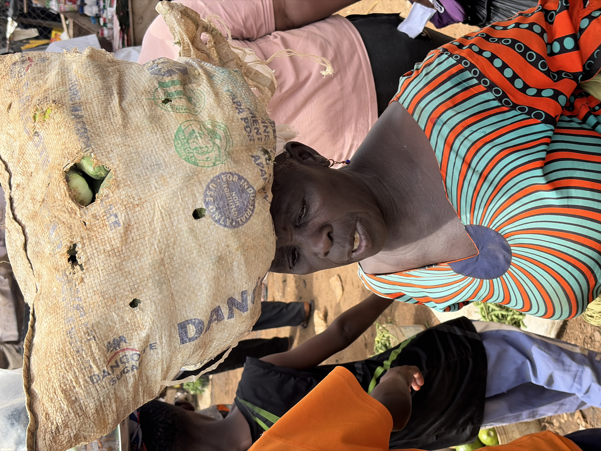 In Gura Top, Nigeria, a woman is seen in a bustling marketplace, carrying a large sack of goods on her head. The sack, made of rough, woven material, appears to be for industrial use, as indicated by the faint text and logos visible on its surface, including "DANGOTE SUGAR". Several gourds or similar produce are visible peeking through a hole in the sack. The woman is wearing a brightly colored, patterned dress with swirling lines of orange and teal. Her expression is focused and her mouth is slightly open, suggesting she might be speaking or reacting to her surroundings. Behind her, other individuals and stalls indicate the active nature of the market. The ground is a mixture of dirt and sand, typical of an outdoor market setting.