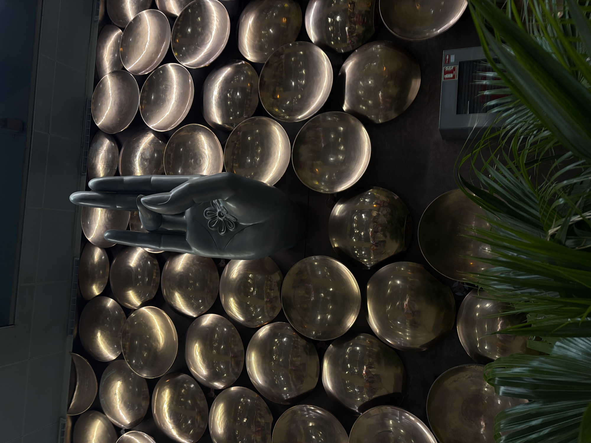 This is a medium shot, eye-level, outdoor, full color, high-angle, dimly lit, eye-level photograph of a large, dark gray sculpture of a hand, with two fingers extended upward in a gesture. The sculpture is positioned in front of a wall decorated with numerous round, metallic, concave discs. In the lower right corner of the frame, a gray metal fire extinguisher cabinet with a red "Fire Extinguisher" label is visible. Lush green palm fronds are in the foreground, partially obscuring the lower portion of the wall and the fire extinguisher. The background consists of a dark ceiling with what appears to be overhead lighting. The style of the hand sculpture and the metallic disc wall suggest a modern artistic installation. This artwork is located in Terminal 3 of the Indira Gandhi International Airport in Delhi, India, as part of its public art collection, designed to evoke spiritual and cultural themes.