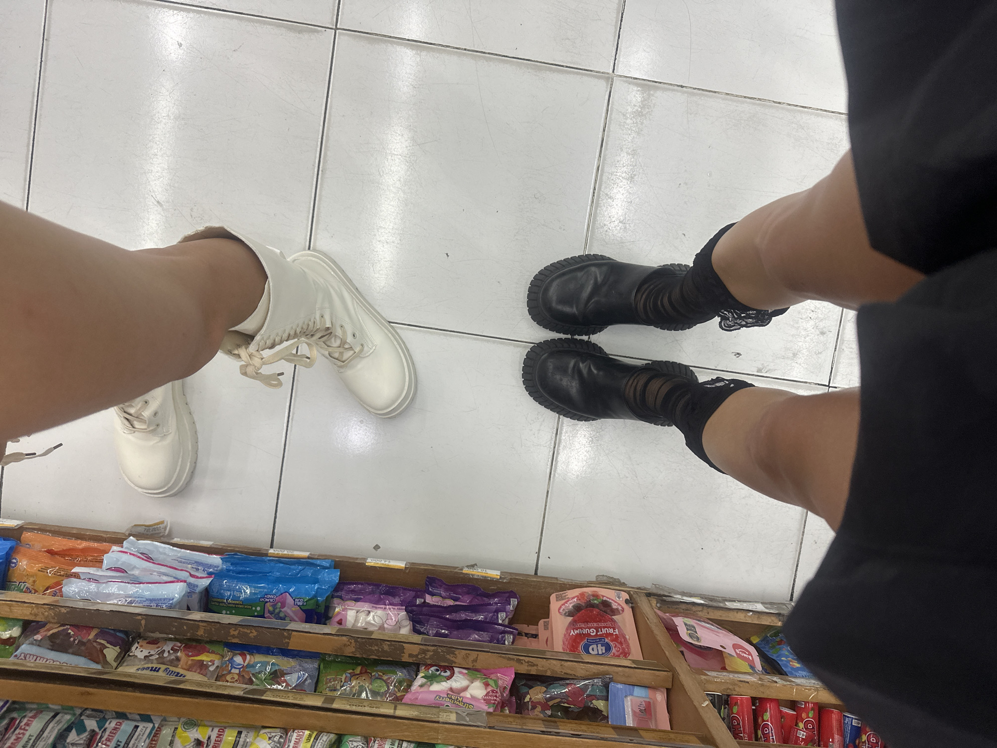 The image depicts a close-up view of two individuals standing in front of a shelf filled with snacks and candies. The location appears to be a store in North Kuta, Indonesia. One person is wearing white platform sneakers, while the other is wearing black boots with striped socks. The snack shelf contains various products, including chips, candies, and gum. The wooden shelf is labeled with prices and brand names. The flooring consists of white tiles.