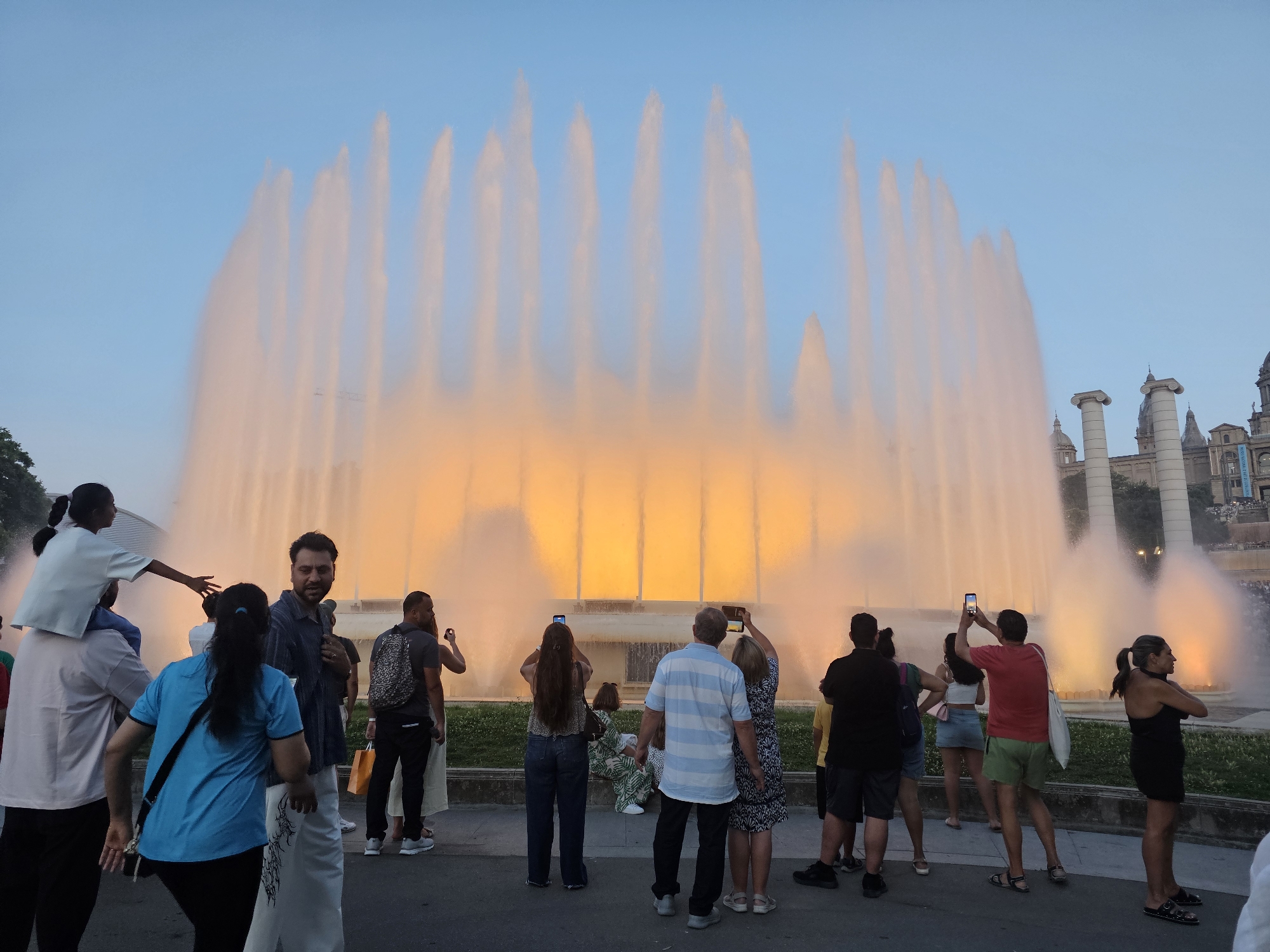 This media file depicts a large crowd gathered to watch a spectacular water fountain show in front of a grand architectural structure. The fountain is illuminated with warm, orange and yellow lights, creating a dramatic effect as water streams upwards against a clear, pale blue sky. The time of day appears to be late afternoon or early evening, as indicated by the soft lighting and the presence of artificial illumination on the fountain.
Several people are visible in the foreground and mid-ground, many of whom are holding up their phones to capture the spectacle. Among the crowd, there are individuals of various ages, dressed in casual summer attire. One child is being carried on another person's shoulders, looking towards the fountain. The overall atmosphere seems to be one of awe and enjoyment.
In the background, the architecture hints at a significant landmark. Two large classical columns stand prominently, with domes and spires of other buildings visible behind them. This architectural style, combined with the context of Barcelona, Spain, suggests the location is likely the Magic Fountain of Montjuïc, situated near the Palau Nacional. The setting is an open area with some greenery and paved walkways, typical of a public plaza or park.
There is no visible text in the image that can be read clearly. The emotion conveyed is one of shared experience and appreciation for the visual display.