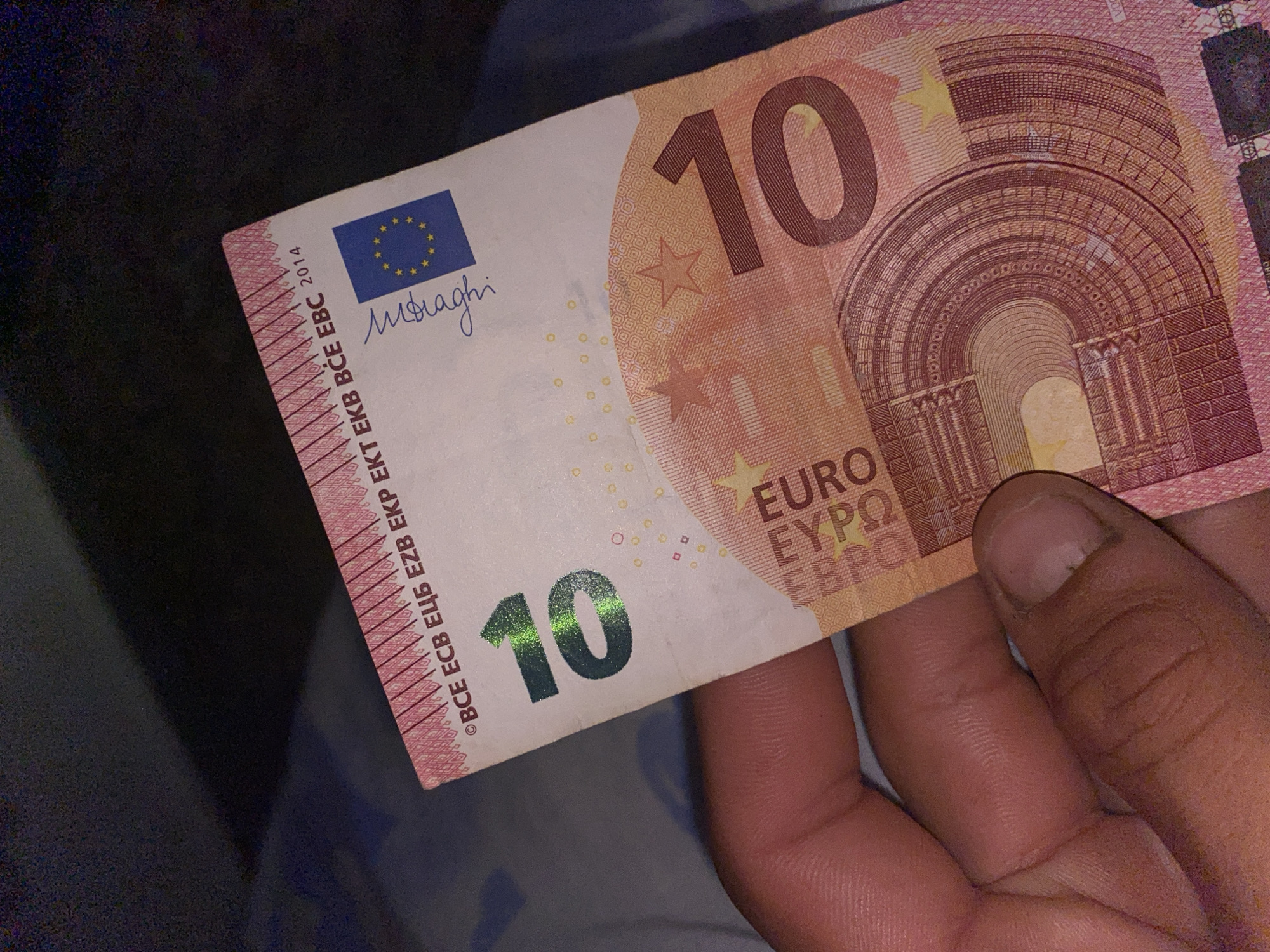 The image shows a close-up of a 10 Euro banknote held by a person's hand. The banknote is light pink and features the number '10' prominently in green, with a holographic shimmer. The European Union flag is visible on the right side, along with a signature that appears to be 'M. Draghi'. Text including 'BCE ECB EZB EKP EKT EKB BCE EBC 2014' is printed along the top edge of the banknote. The background is dark and out of focus. There is no information in the image that can be used to determine the location in the city of Brashlen, Bulgaria.