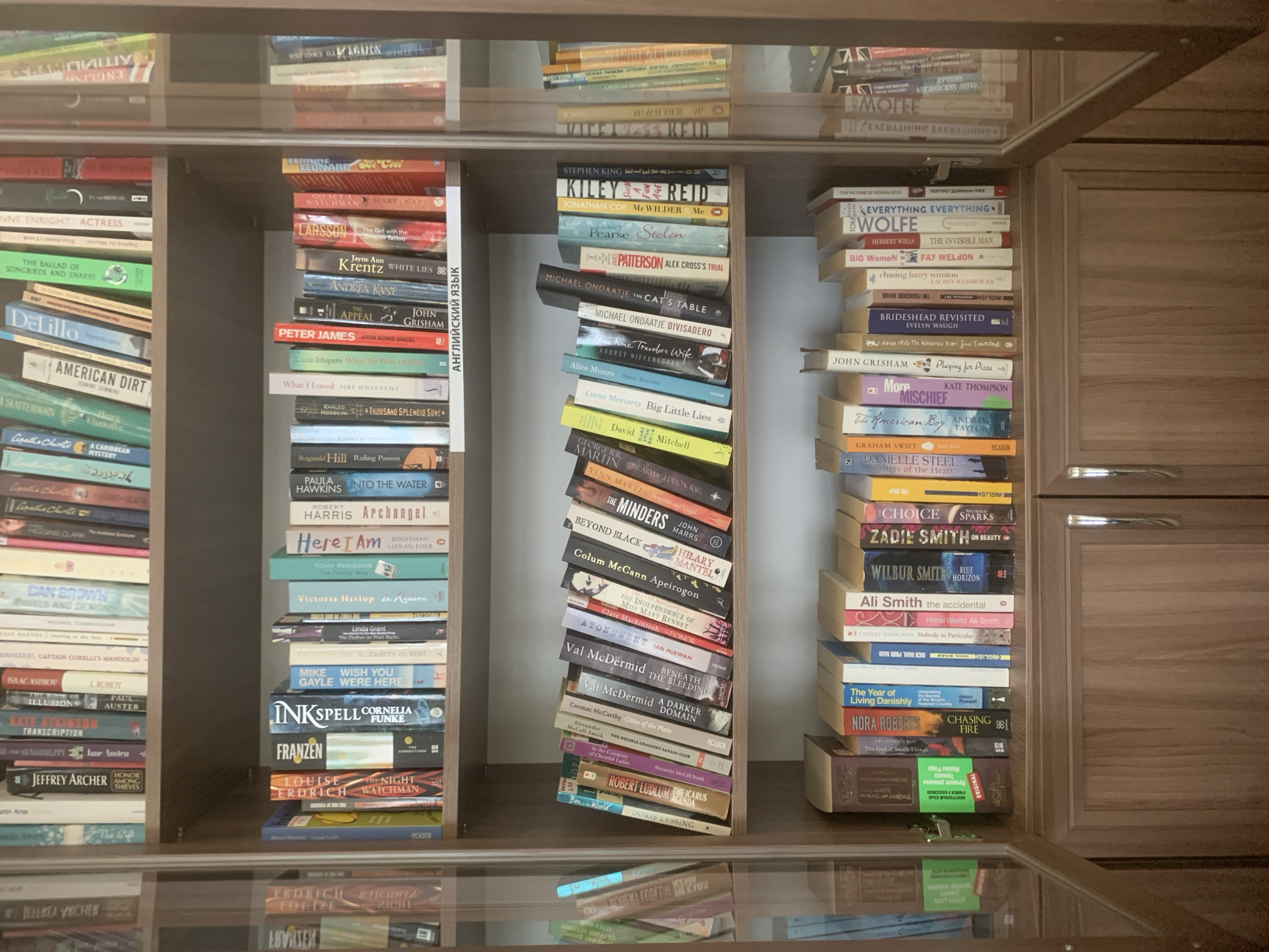 The image shows a bookshelf filled with books. The bookshelf is made of brown wood, with two doors visible on the right side. The books are arranged vertically and horizontally in three columns. The left column contains books with titles such as "American Dirt," "I, Robot," and "Honor Among Thieves." The middle column contains books with titles such as "Mr Wilder Me," "The Cat's Table," "Big Little Lies," "The Minders," and "Atonement." These books are tilted. The right column contains books with titles such as "Everything, Everything," "The Invisible Man," "Brideshead Revisited," "Playing for Pizza," and "The Year of Living Danishly." The books are reflected in a glass surface at the bottom of the image. The image was not taken in Izhevsk, Russia, the city is not visible. There are no people in the image.