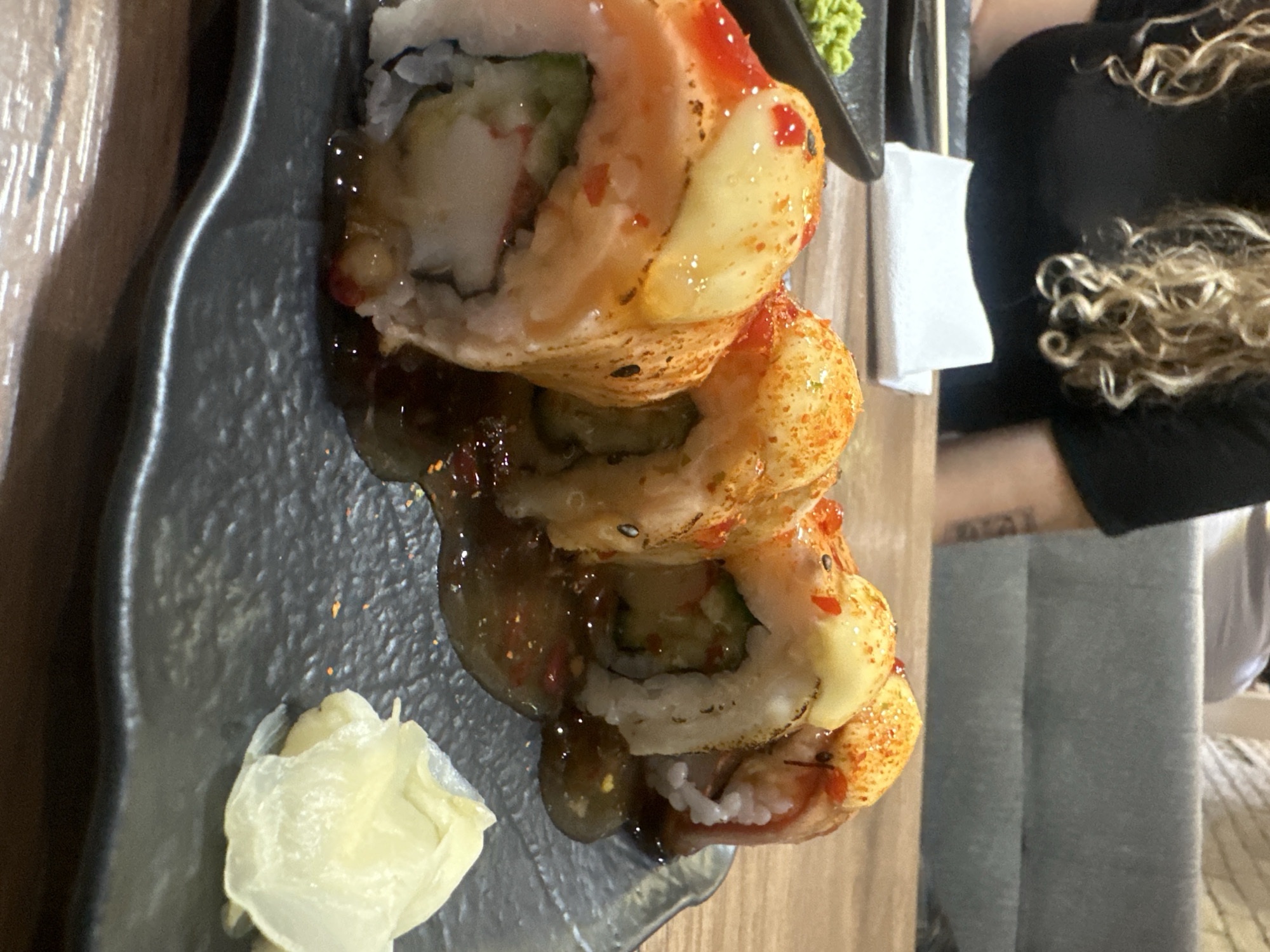 A close-up, vertically-oriented photograph shows a plate of sushi. The sushi is stacked in a tower of three pieces, covered with a sauce and a red and yellow topping. The sushi appears to be drizzled with a dark sauce. Next to the sushi is a small pile of what looks like pickled ginger. The plate is dark gray and has a slightly ornate edge.
The photograph is taken from a low angle, looking up at the sushi. The background is blurry, but it appears to be a wooden table with a person sitting to the right. The person has blonde curly hair, is wearing a black shirt, and gray pants. The location is unidentifiable as Varna, Bulgaria.