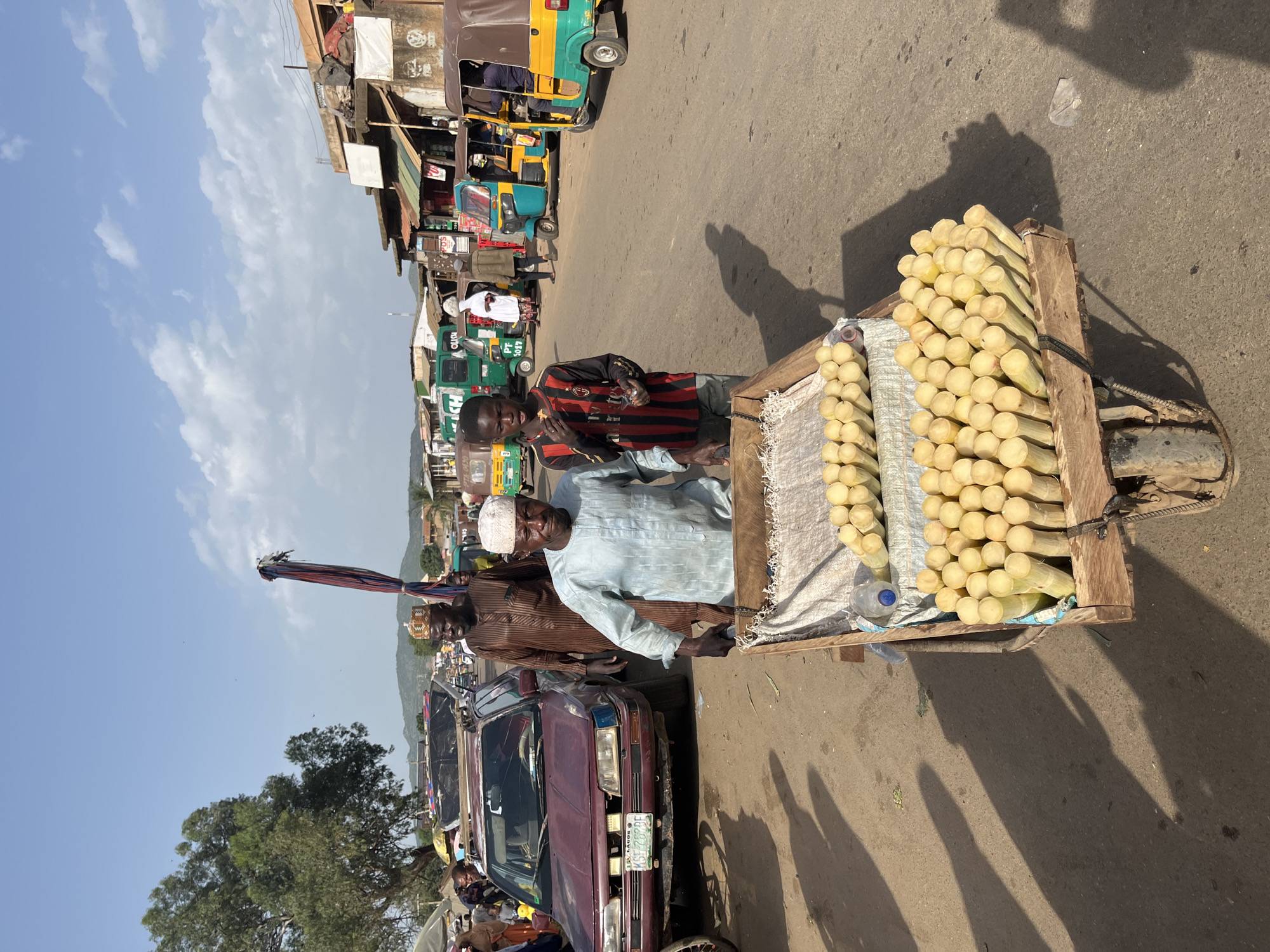 In the city of Rafin Par, Nigeria, a street scene unfolds under a bright, partly cloudy sky. The foreground is dominated by a wooden cart filled with stalks of sugarcane. Two men are present; one, wearing a light blue tunic and white cap, stands behind the cart, appearing to be a vendor. Another man, dressed in a brown traditional robe and a beaded cap, stands to the left of the cart. A young boy in a striped shirt is positioned to the left of the vendor, looking towards the camera and holding a piece of sugarcane.
Behind them, a maroon sedan is parked on the dirt road. Further back, a bustling marketplace is visible, with various stalls and shops lining the street. Several auto-rickshaws, brightly colored in yellow, green, and blue, are also part of the scene, suggesting a busy transportation hub. The background reveals a line of buildings and a hilly landscape. The overall impression is one of a lively and active marketplace in a Nigerian town.