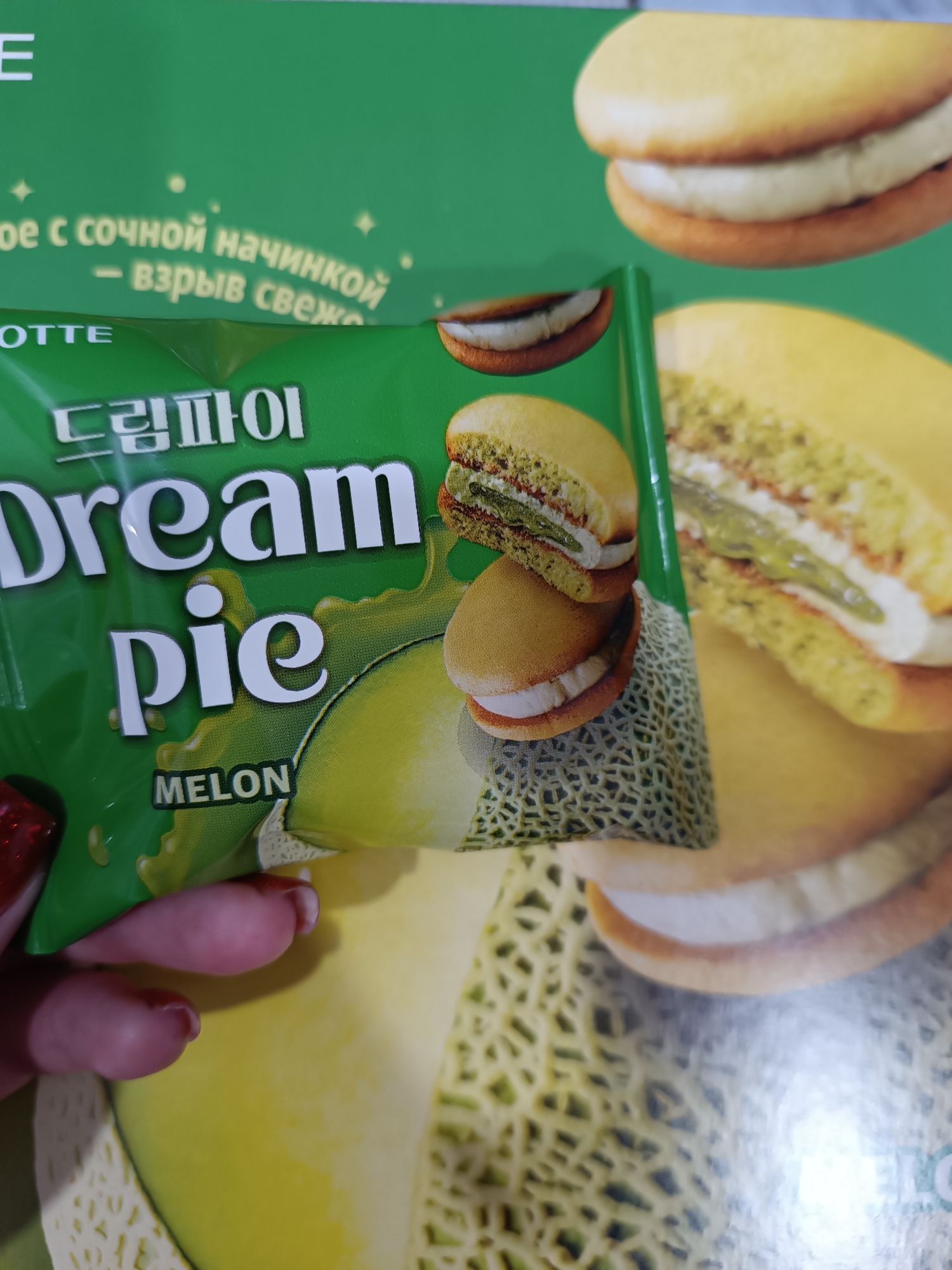 A hand with red nail polish holds a green package of "Dream Pie" melon-flavored snacks. The packaging displays an image of a melon and two cookies stacked, revealing a filling between them. The backdrop consists of similar larger images of the same cookies and melons. The text on the package is in English and Korean. The photo appears to be taken indoors. I have no information to determine the location of this image.