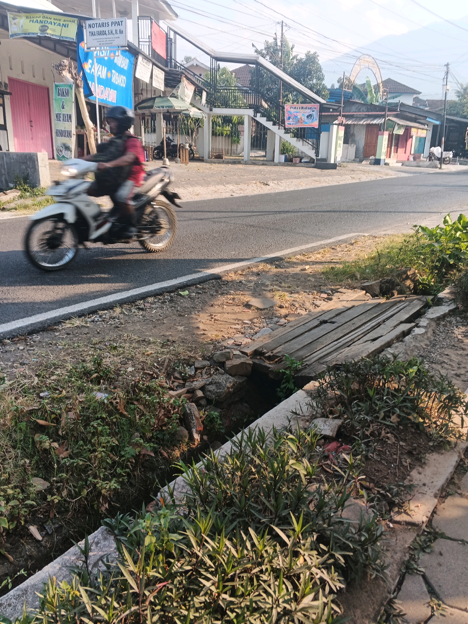 The image depicts a street scene in Ungaran, Indonesia. In the foreground, a drainage ditch runs along the side of the road, covered by a makeshift wooden bridge. Lush green vegetation grows around the ditch and on the embankment. A motorcycle rider wearing a helmet and a dark vest is captured in motion, traveling on the asphalt road. In the background, a row of buildings lines the street, featuring shops with various signs, including "NOTARIS PPAT TIKA FARIDA, S.H., M.Kn" and "BAKSO DAN MIE AYAM HANDAYANI." A pedestrian bridge can be seen further down the street, and an arched structure is visible in the distance. The sky is clear, with mountains visible in the background, suggesting a rural or semi-urban setting. The lighting indicates it is daytime.