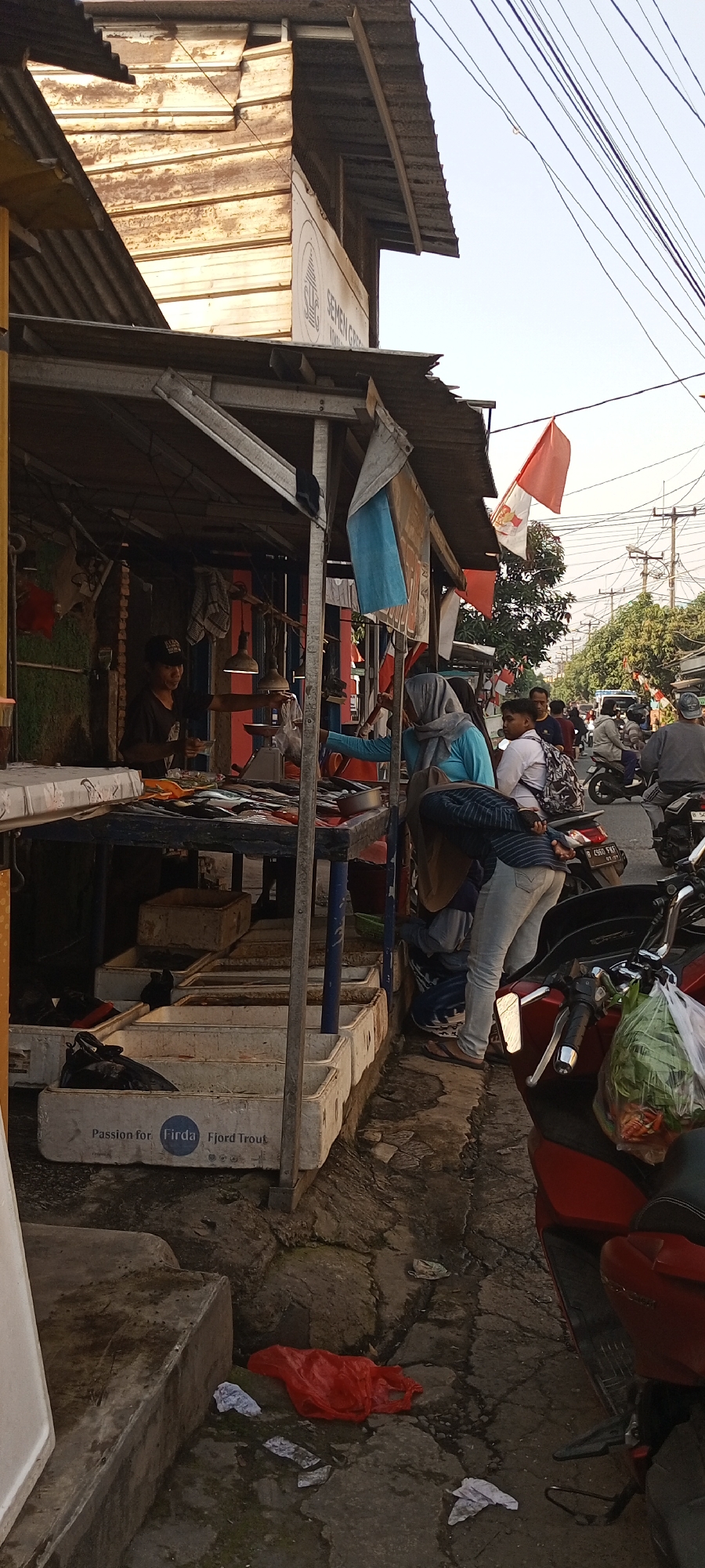 The image depicts an outdoor market scene on a sunny day, likely in Setu, Indonesia. In the foreground, a red motorcycle is parked with a plastic bag of vegetables on its seat. The ground is paved with rough stones and scattered with debris, including plastic bags and wrappers.
The central area of the image features a market stall with a corrugated metal roof supported by metal poles. Inside the stall, a man wearing a black t-shirt and a baseball cap stands behind a table displaying fish on ice. A mechanical scale is also visible. In front of the stall, several white plastic crates are arranged, some containing fish. One crate has the text "Passion for. Firda. Fjord Trout" printed on it.
Several people are present. A woman in a light blue headscarf and long-sleeved shirt is interacting with someone at the stall, possibly making a purchase. Another person, wearing jeans and a light-colored shirt, is bending down near the stall. Further in the background, several other individuals are visible, some on motorcycles. One motorcycle has a license plate that reads "B 4860 FWF". In the background, there are more market stalls and trees, with power lines crisscrossing the sky. A red and white Indonesian flag is flying from a pole above one of the stalls. The overall impression is one of a bustling, open-air market.