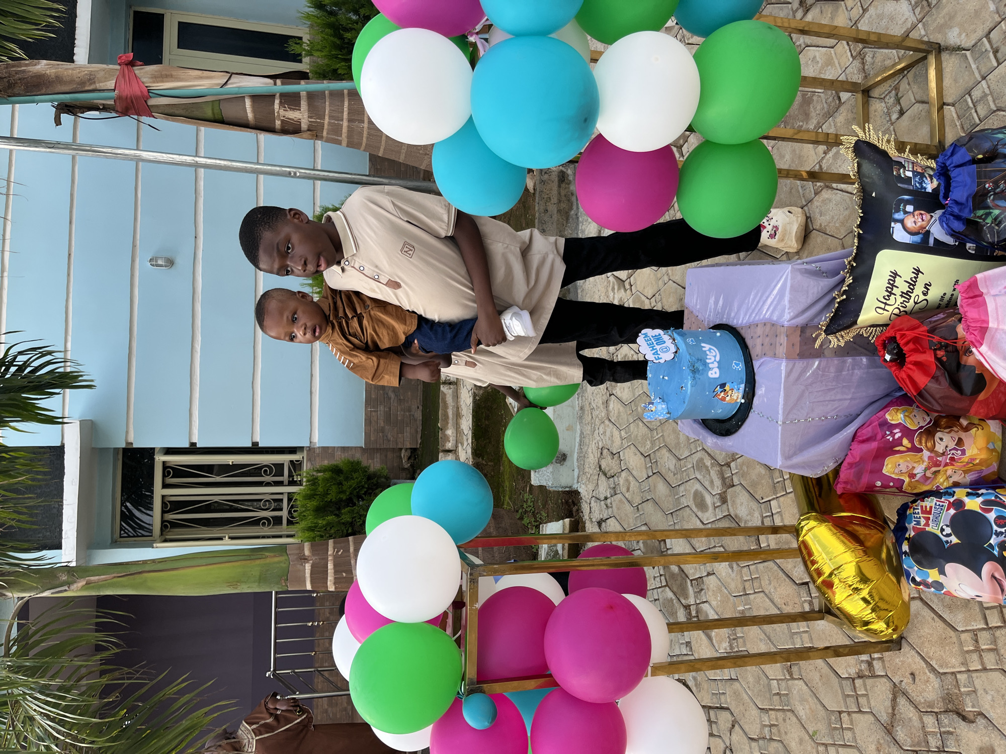 This full-frame image depicts an outdoor birthday celebration in the city of Jos, Nigeria, featuring two male children positioned centrally within a festive setup.
The older child, on the right, is a boy with short dark hair, wearing a light beige collared polo shirt with a dark brown collar and button placket, and dark long trousers. He is holding a younger child, a toddler, who is dressed in a brown long-sleeved shirt with subtle embroidery, blue jeans, and white sneakers. The toddler faces left, looking forward, while the older boy looks slightly to his right with his mouth slightly ajar.
The scene is adorned with clusters of balloons in green, white, pink, and turquoise, arranged vertically on two gold-colored metallic stands flanking the children. In front of the children, on a small table draped in light purple fabric, sits a light blue, two-tier round birthday cake. The top tier is inscribed with "FAHEEM @ ONE" in white lettering, and the bottom tier features "Bluey" in white lettering alongside a character illustration, topped with a small Bluey figure.
On the paved ground surrounding the table are various party favors and gifts. These include a deflated gold foil balloon, and several drawstring bags: one featuring Mickey Mouse with the text "MEET ME IN THE CLUBHOUSE!", another depicting Disney Princesses, a red bag, and a partially visible blue bag with a character design. A black decorative cushion with gold fringe, bearing the text "Happy Birthday Son" and a photograph of a smiling child, rests among the bags.
The background features a light blue painted residential building with horizontal structural ridges and windows fitted with white security bars. A tall palm tree stands prominently to the right of the building, with another partially visible on the far left. Green shrubbery is present near the building's foundation. On the far left, partially obscured by the palm tree, an individual wearing a brown patterned garment is visible. The ground is covered with light-colored interlocking pavers. The scene is illuminated by natural daylight.