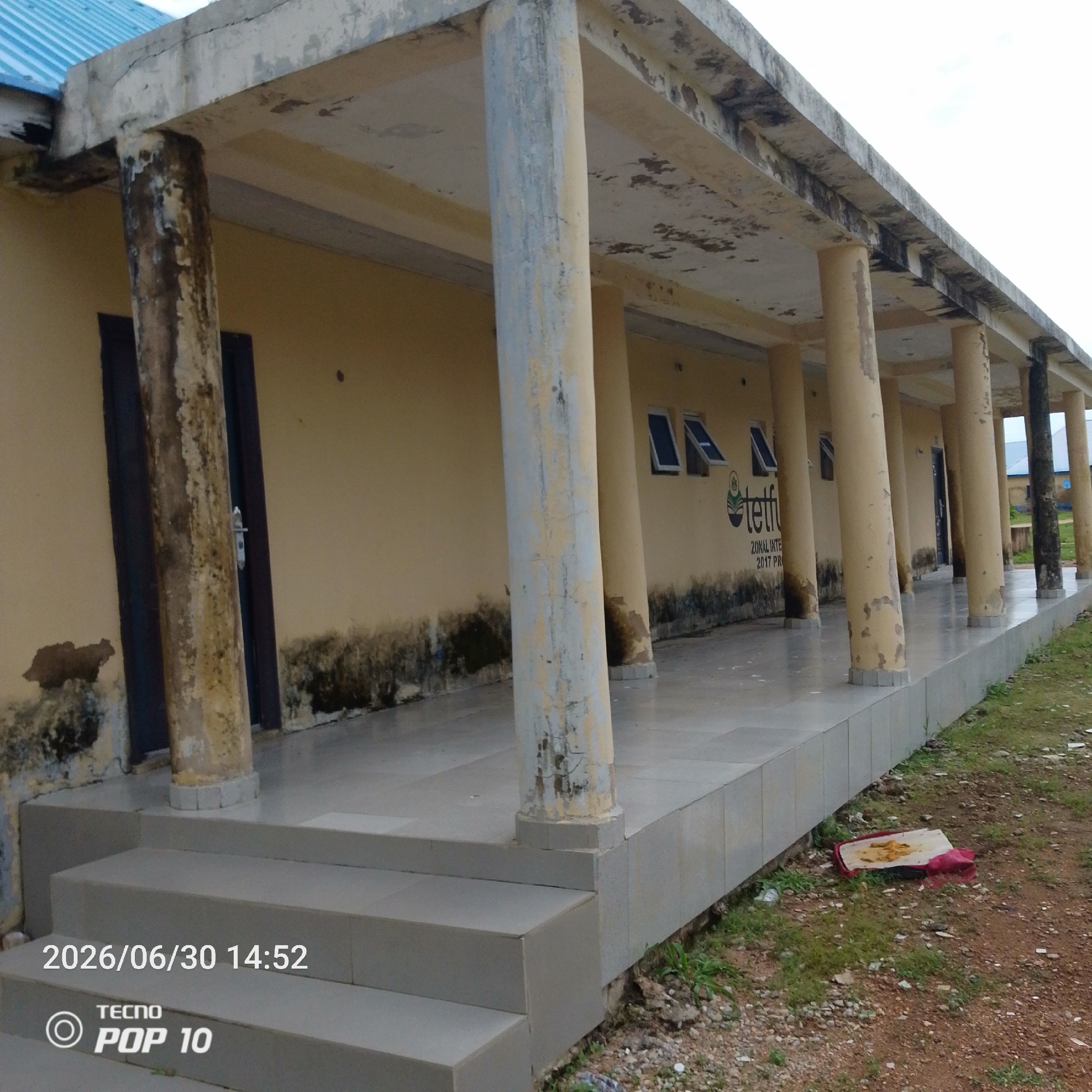 The image depicts the exterior of a building with a covered walkway supported by pillars. The building's walls are painted a pale yellow and show signs of wear, with dark stains and peeling paint, particularly at the base. The walkway has a tiled floor and ceiling, which also exhibits some discoloration and damage. Several windows, some open, are visible on the building's wall. A logo with text that includes "tetfu" and "ZONAL INTE 2017 PR" is present on the wall. The walkway leads to steps that descend to ground level. The ground around the building is covered in rough terrain with grass, stones, and debris, including a red object with some yellow material on it. The sky is overcast. The timestamp on the image indicates June 30, 2026, at 14:52, and the phone model is TECNO POP 10. The location is identified as Jalingo, Nigeria. There are no people visible in the image.