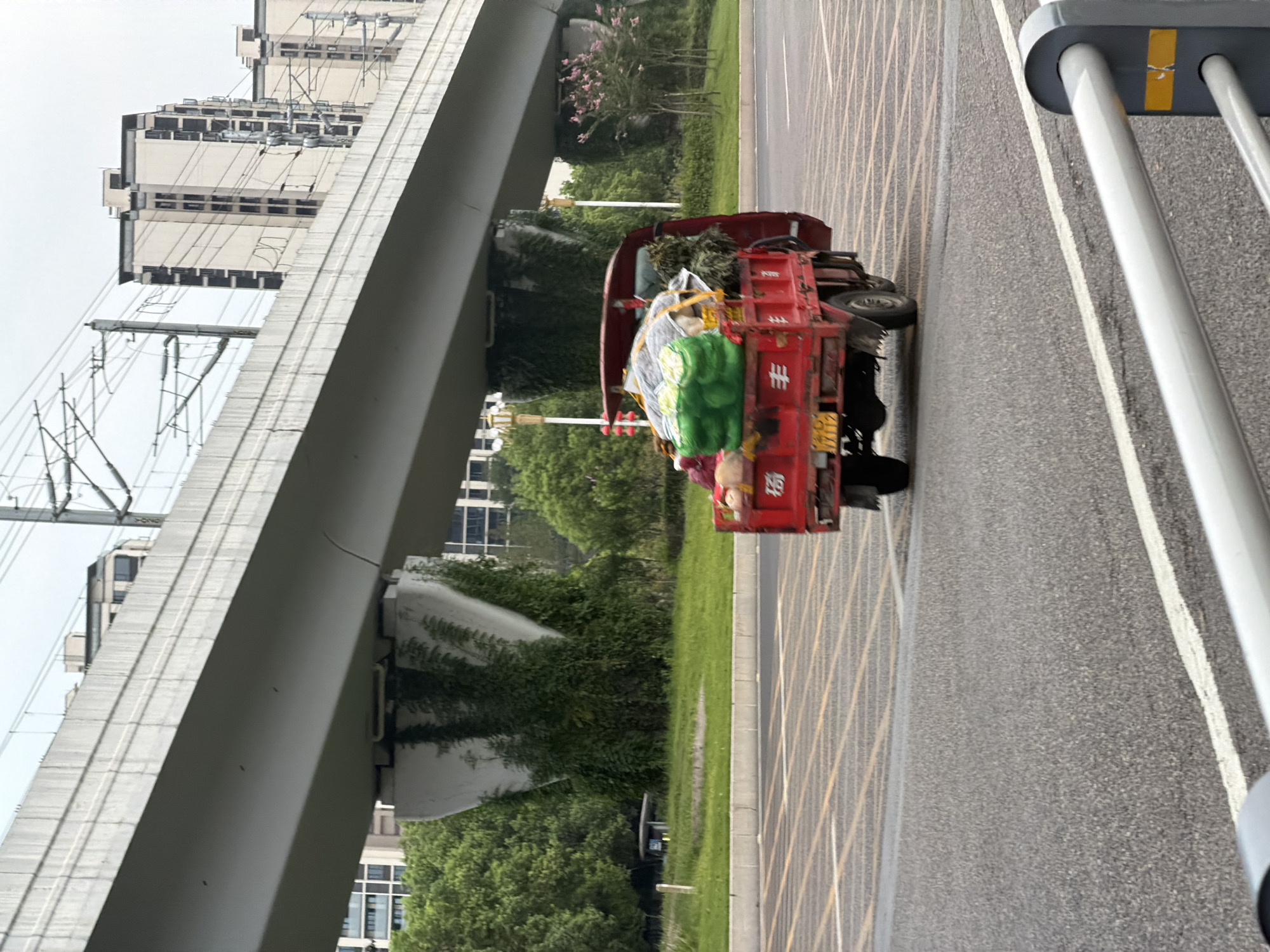 In Jiangjin, China, a red truck is driving under an elevated highway overpass. The truck is carrying a load of what appears to be vegetables wrapped in green netting, as well as some other items. The truck is red and has a yellow license plate. The highway overpass is concrete and has electrical wires running above it. There are trees and buildings in the background. The road is asphalt and has yellow and white lane markings.