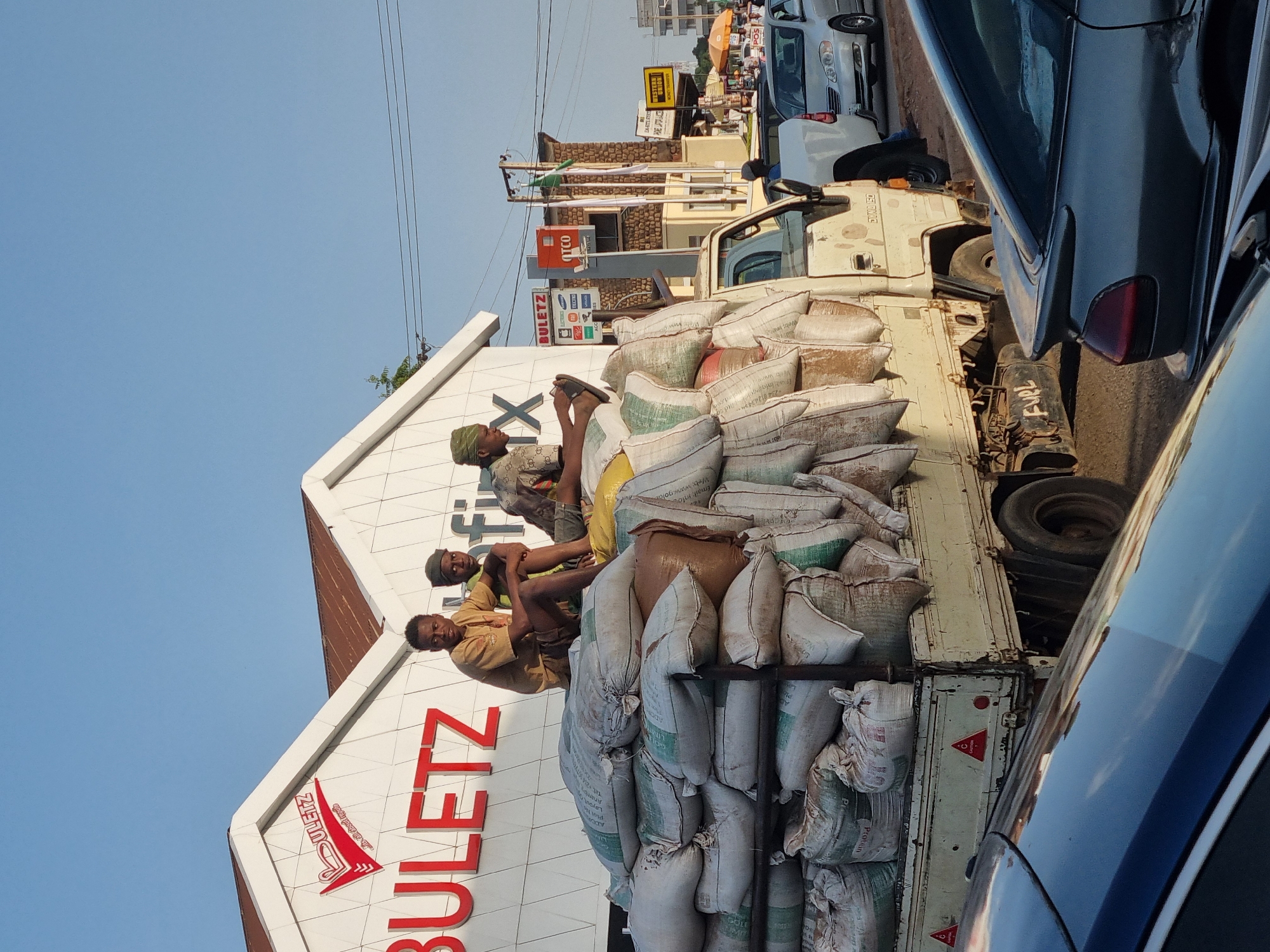Three men are seated atop a truck overloaded with sacks, likely carrying goods or produce. One man is wearing a green hat. They are in front of a building with a large "BULETZ" sign, in front of DYNA 150. Another sign is of Western Union. The scene appears to be in Jos, Nigeria, identified by the "Anamba" sign which is an address of Jos. The truck is parked on a street with other vehicles nearby. The weather is clear.