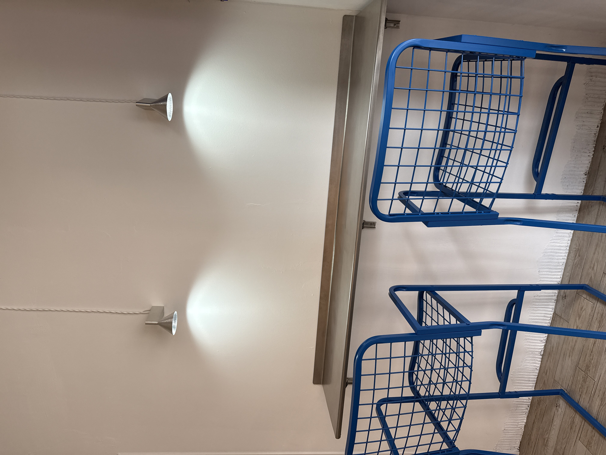 The image shows a room with two blue metal chairs stacked next to a shelf. Two overhead lights are mounted on the white wall, casting a bright spotlight. The background suggests a modern interior setting. The location of the image cannot be determined as Krasnoyarsk, Russia, from the provided image alone.