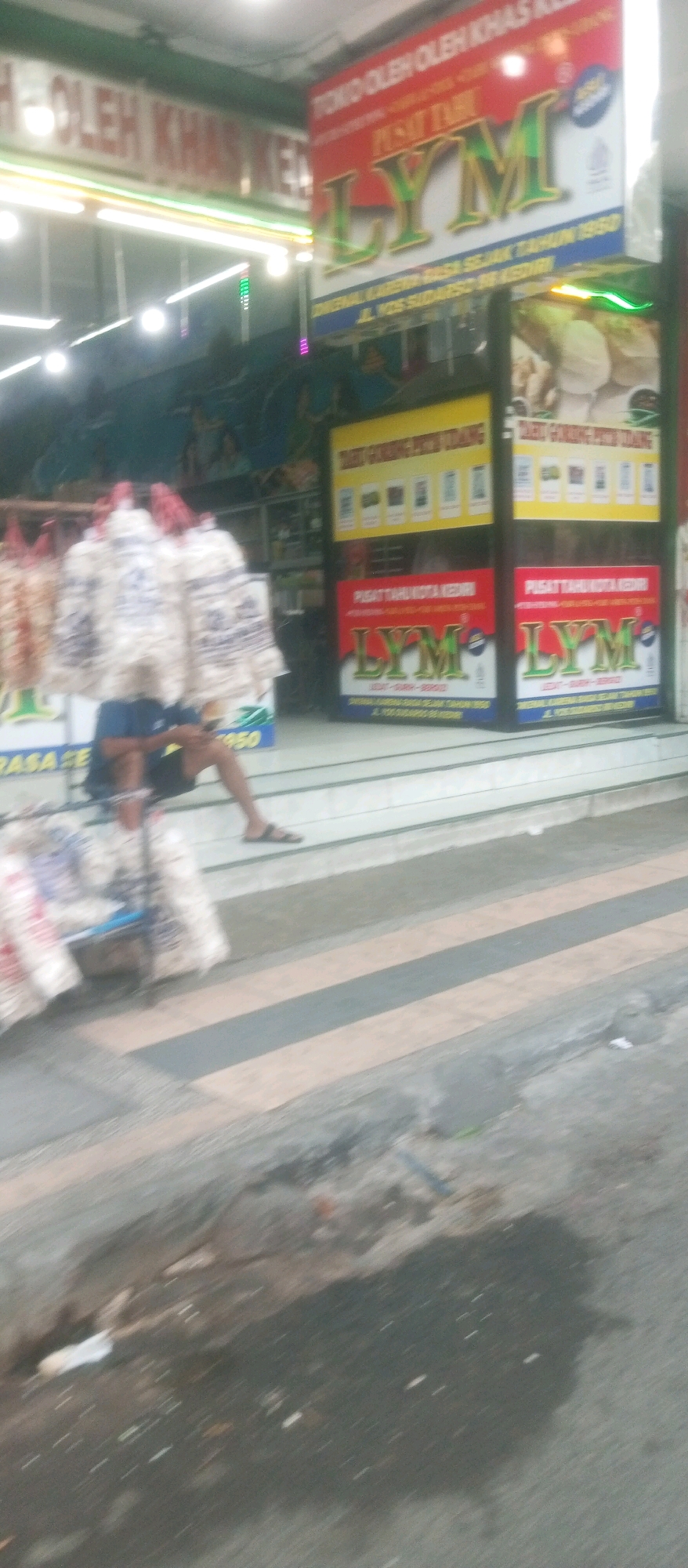 The image depicts a blurred street scene in front of a shop. The shop has signage that reads "TOKO OLEH OLEH KHAS KEDIRI" and features a prominent logo with the text "LYM". Below this, there are signs indicating "PUSAT TAHU KOTA KEDIRI" and product advertisements. A person is sitting on steps in front of the shop, appearing to be using a mobile phone. Several bags of what appear to be snacks or goods are displayed on a cart to the left of the person. The street in front of the shop is paved with diagonal stripes of light and dark gray. The environment appears to be a commercial area in the city of Kediri, Indonesia, given the signage.