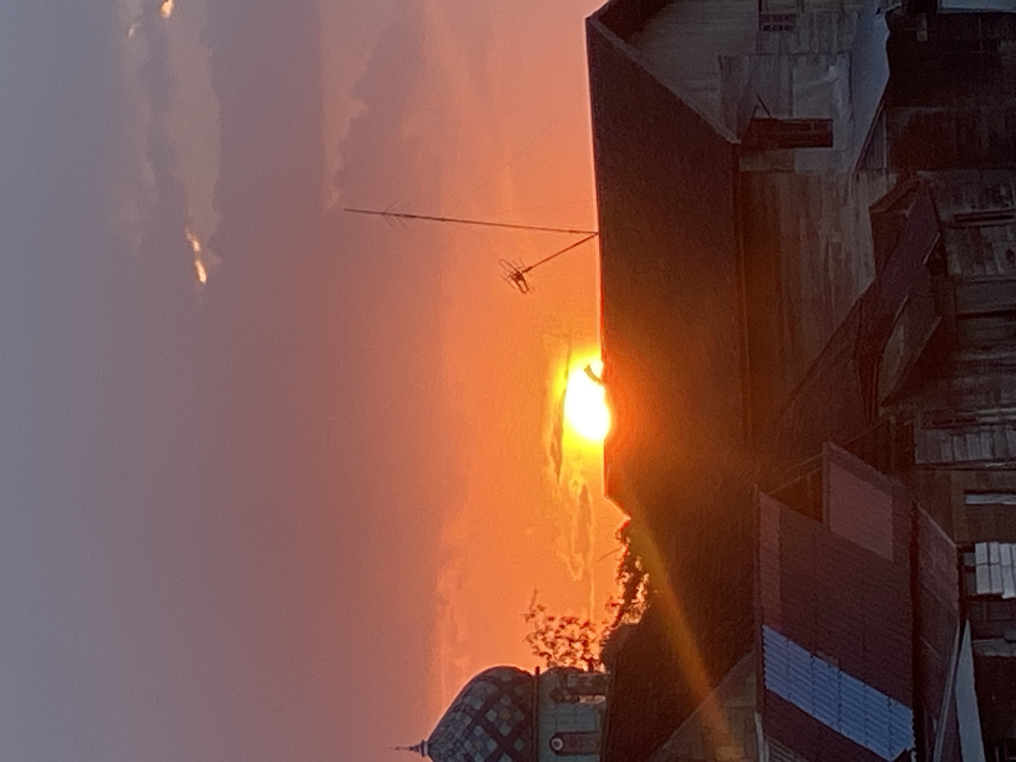 A sunset scene is depicted over the city of Tunggul Irang Ulu, Indonesia. The sky is ablaze with vibrant orange and pink hues, transitioning to a soft purple at the top. The sun, a brilliant orange disc, is partially obscured by the rooftops of several buildings, creating a dramatic silhouette. One prominent building features a dome-shaped roof adorned with intricate patterns and a finial. Other structures appear to be houses or shops with corrugated metal roofs, some showing signs of wear. An antenna stands tall on one of the rooftops, silhouetted against the sky. The overall impression is one of a peaceful evening, with the natural beauty of the sunset dominating the urban landscape.