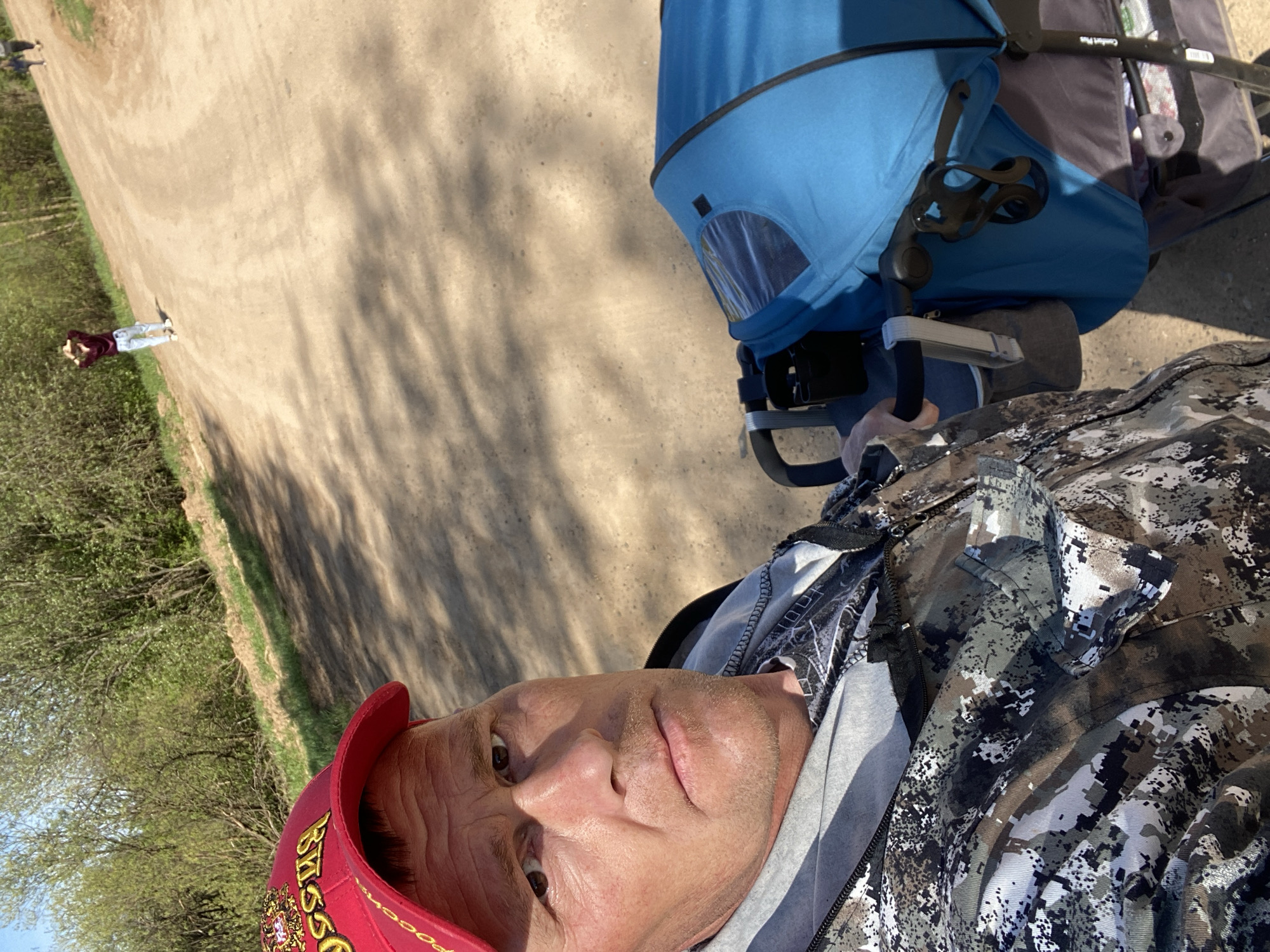 A man wearing a red cap and camouflage jacket is taking a selfie while pushing a blue and gray stroller on a dirt road. In the background, a person walks along the road. The location is Фоки, Russia.
