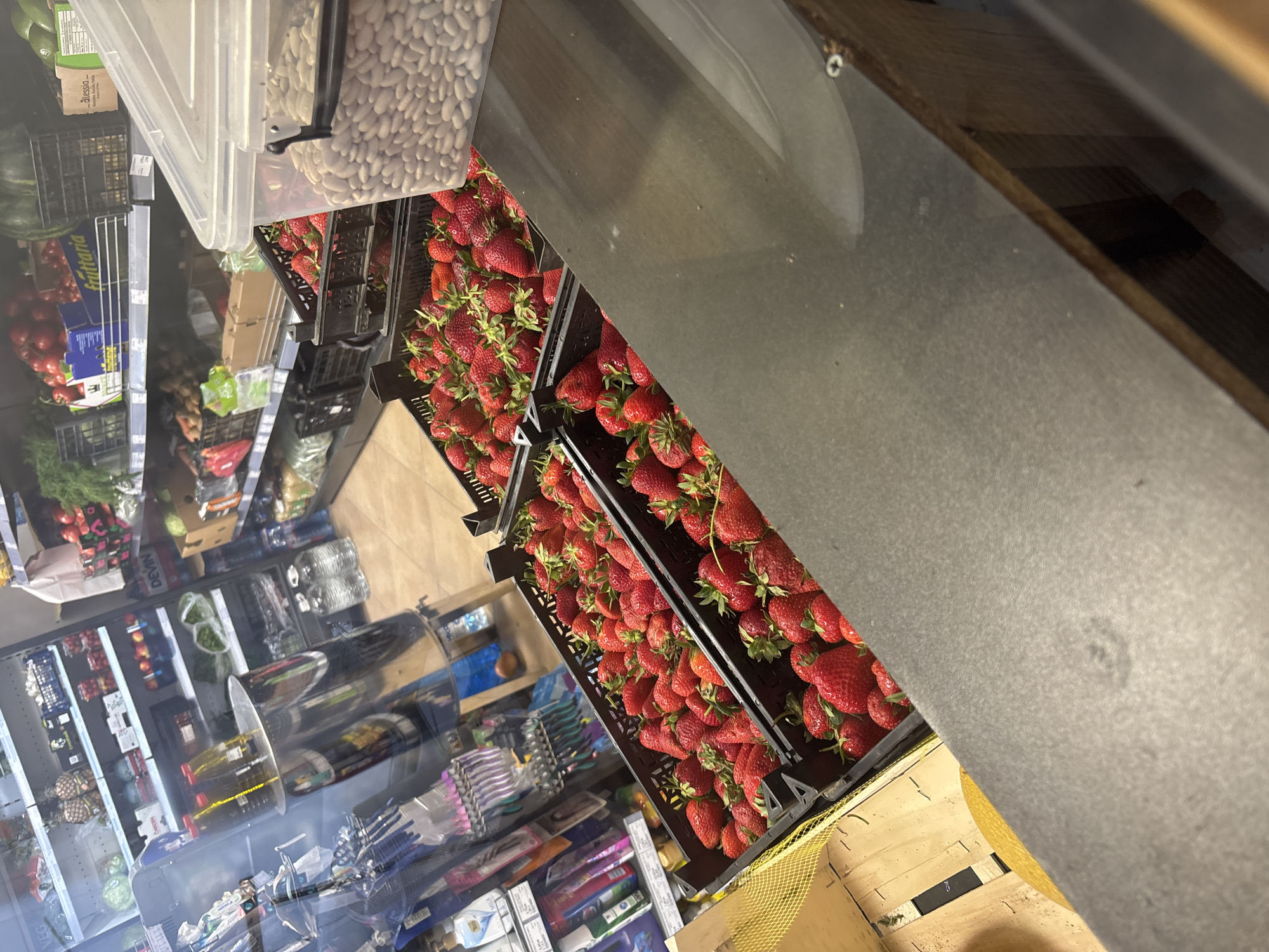 The image depicts a fruit and vegetable display within a store in Plovdiv, Bulgaria. Crates filled with fresh strawberries are prominently featured, stacked in multiple rows. A clear container of white beans sits adjacent to the strawberries. Other produce items, including tomatoes and greens, can be seen in refrigerated display cases in the background. Additional retail products, such as personal care items, are also visible. The lighting appears to be artificial. The angle is a high-angle shot looking down at the fruit, and the image seems to be taken from within the store. The environment is a typical retail setting. No people are visible.